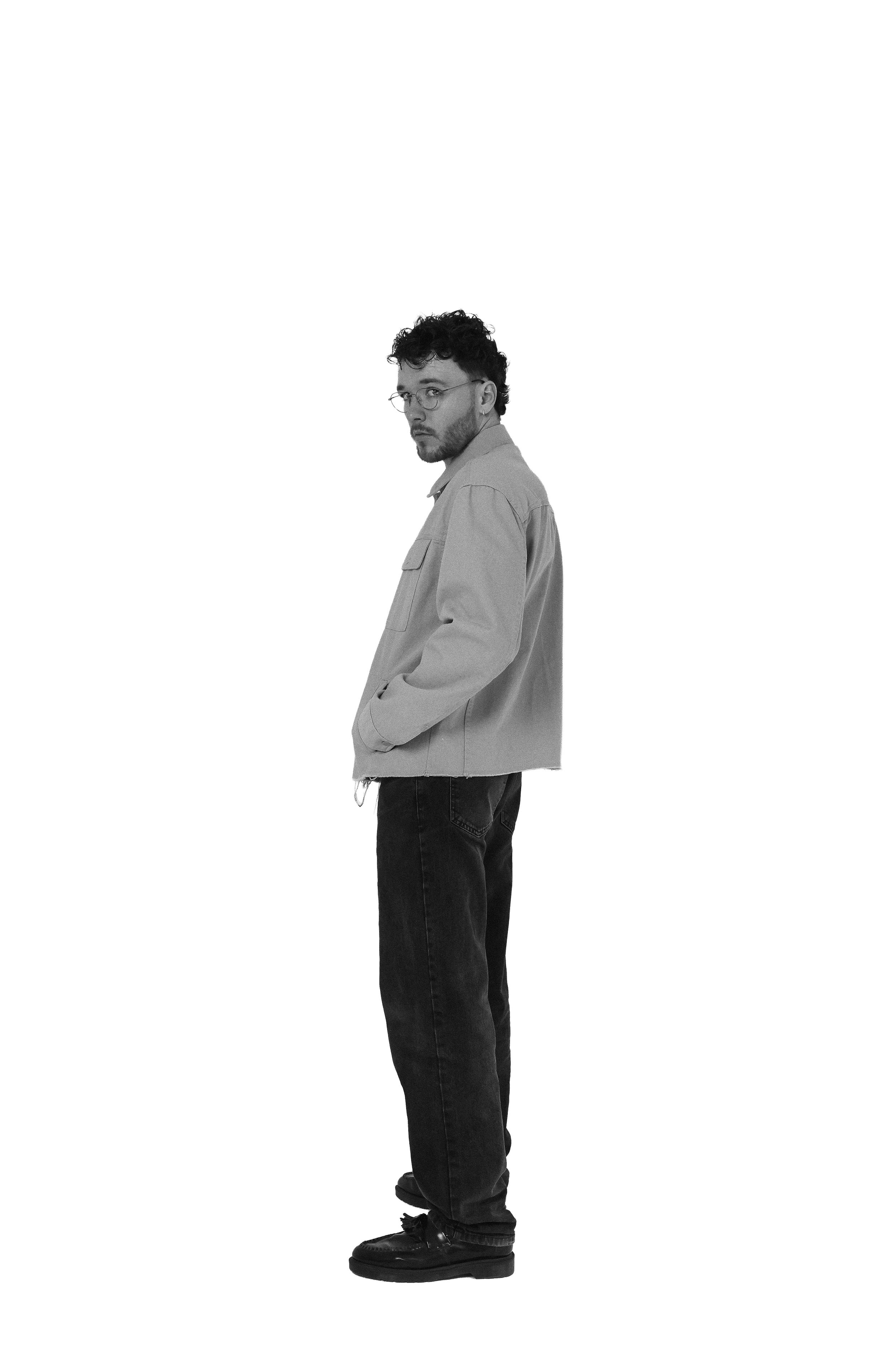 A man with curly dark hair and glasses, wearing a light-colored jacket, black pants, and black shoes, is standing sideways and looking towards the camera in a studio with a plain background.