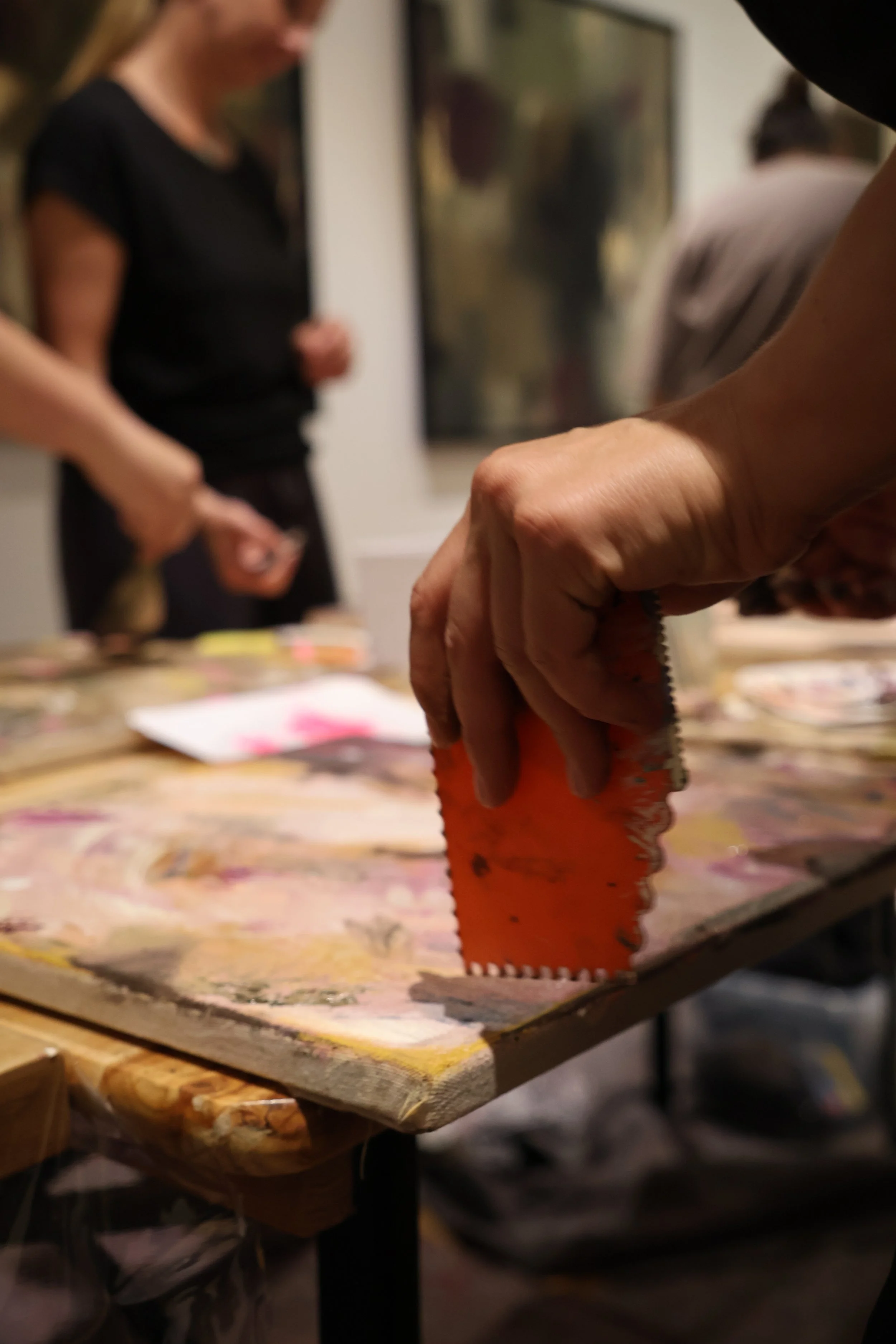Fokus auf eine Hand, die ein Stück orangefarbenes Holz mit einer Säge schneidet. Im Hintergrund sind mehrere Menschen in einem Kunstatelier sichtbar, die an Bildern arbeiten oder zuschauen.