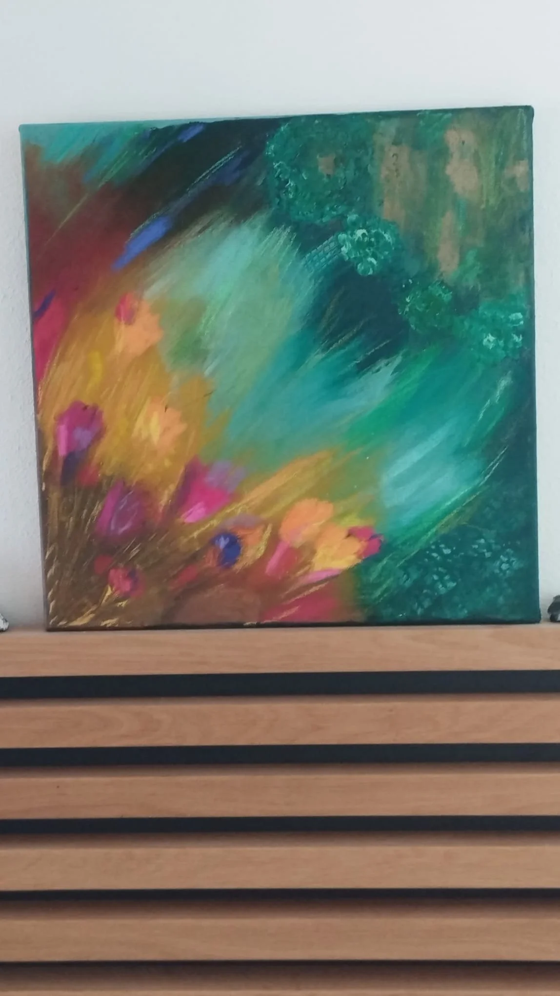 Ein abstraktes Gemälde mit bunten, dynamischen Pinselstrichen in Blau, Grün, Pink und Orange, auf einer weißen Wand und einem Holzregal darunter.