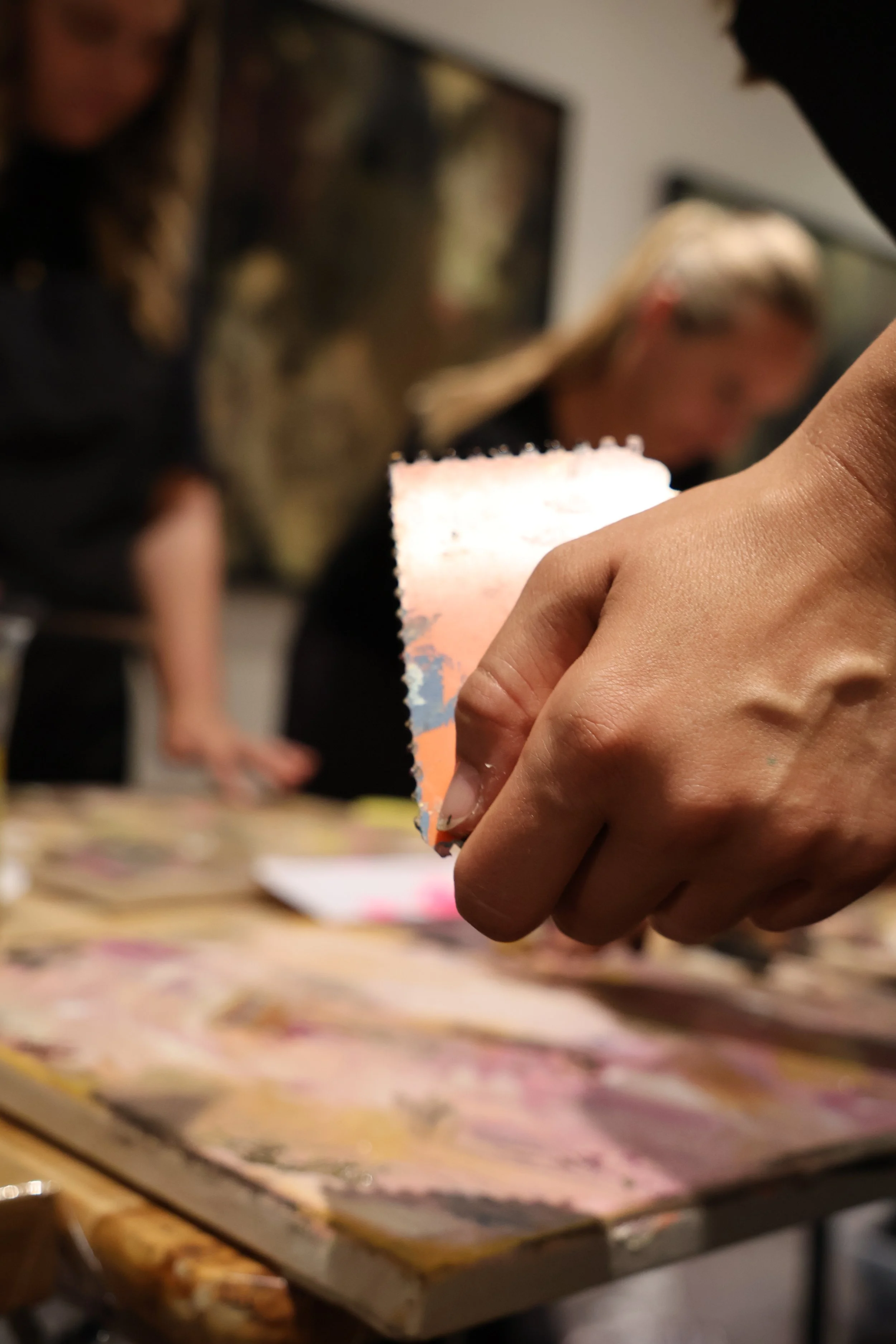 Nahaufnahme einer Hand, die ein zerzaustes Stück Papier hält, im Hintergrund sind zwei Frauen, die an einem Tisch arbeiten, bei einer Aktivität mit buntem Papier.