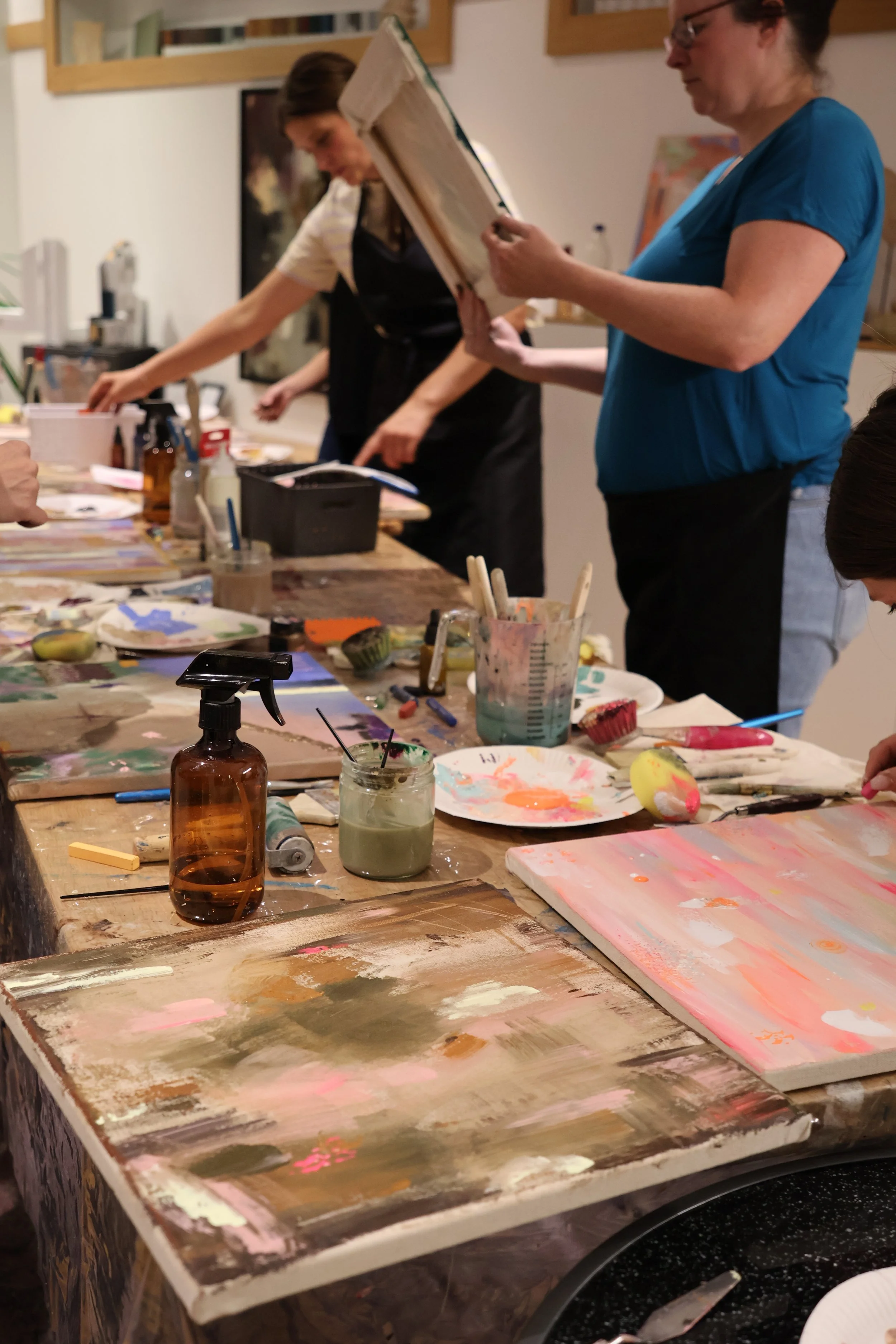Ein Künstleratelier mit mehreren bemalten Leinwänden, Farbpaletten, Pinseln und Malutensilien auf einem Holztisch. Im Hintergrund sind zwei Frauen, die kreative Arbeiten machen.