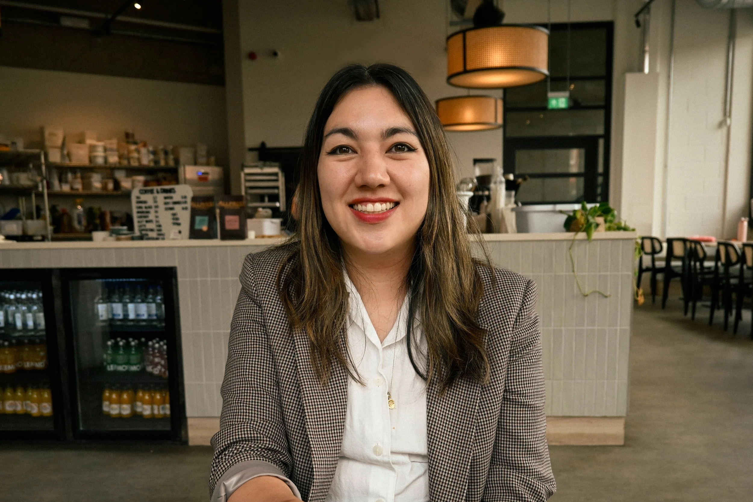 A woman with long brown hair smiling at the camera, sitting in a café or restaurant setting, with shelves of jars and bottles and a menu board in the background.