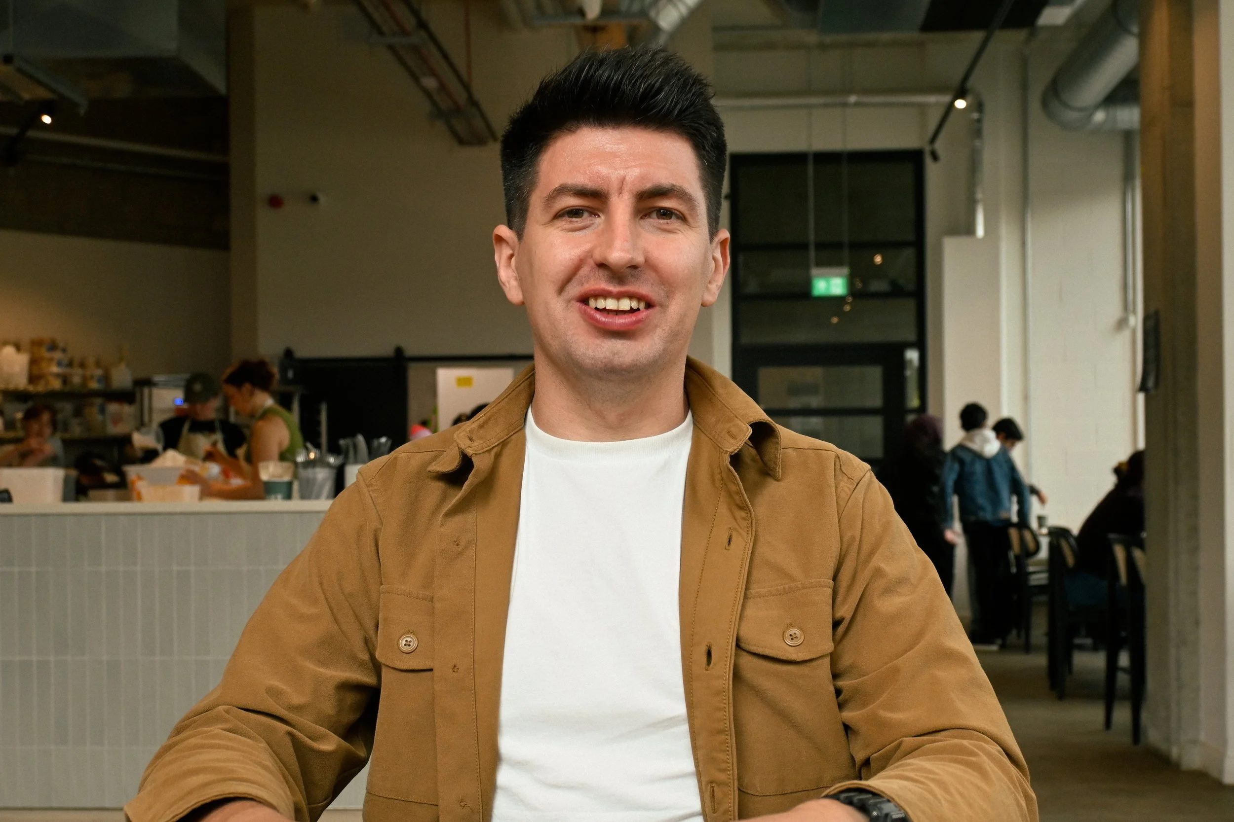 A man with dark hair and light skin is smiling and wearing a brown jacket over a white shirt, sitting in a modern café or restaurant with people and tables in the background.