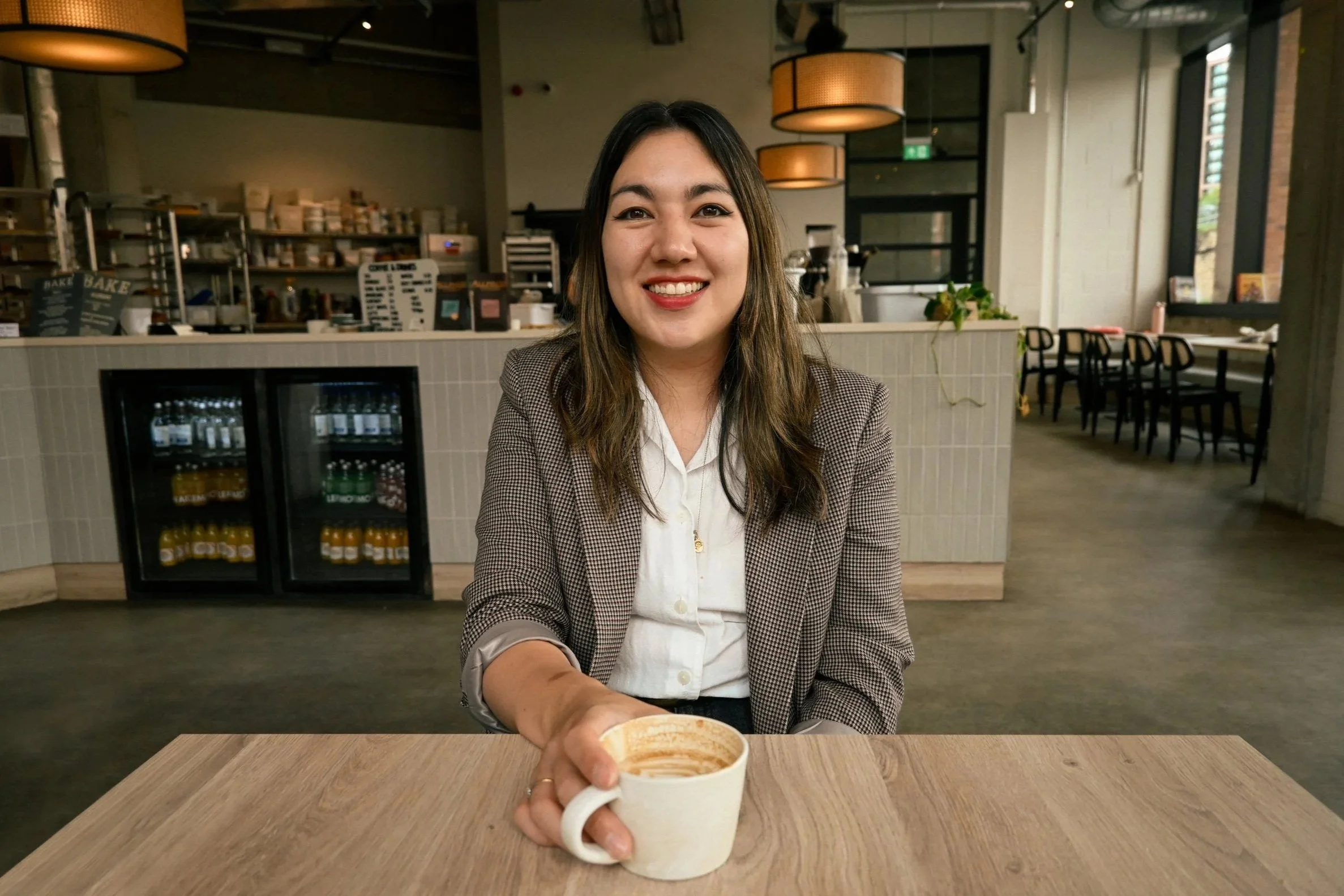 A woman with long brown hair smiling while sitting at a table in a cafe, holding a white mug of coffee, with a background of café interior and a small refrigerator visible.