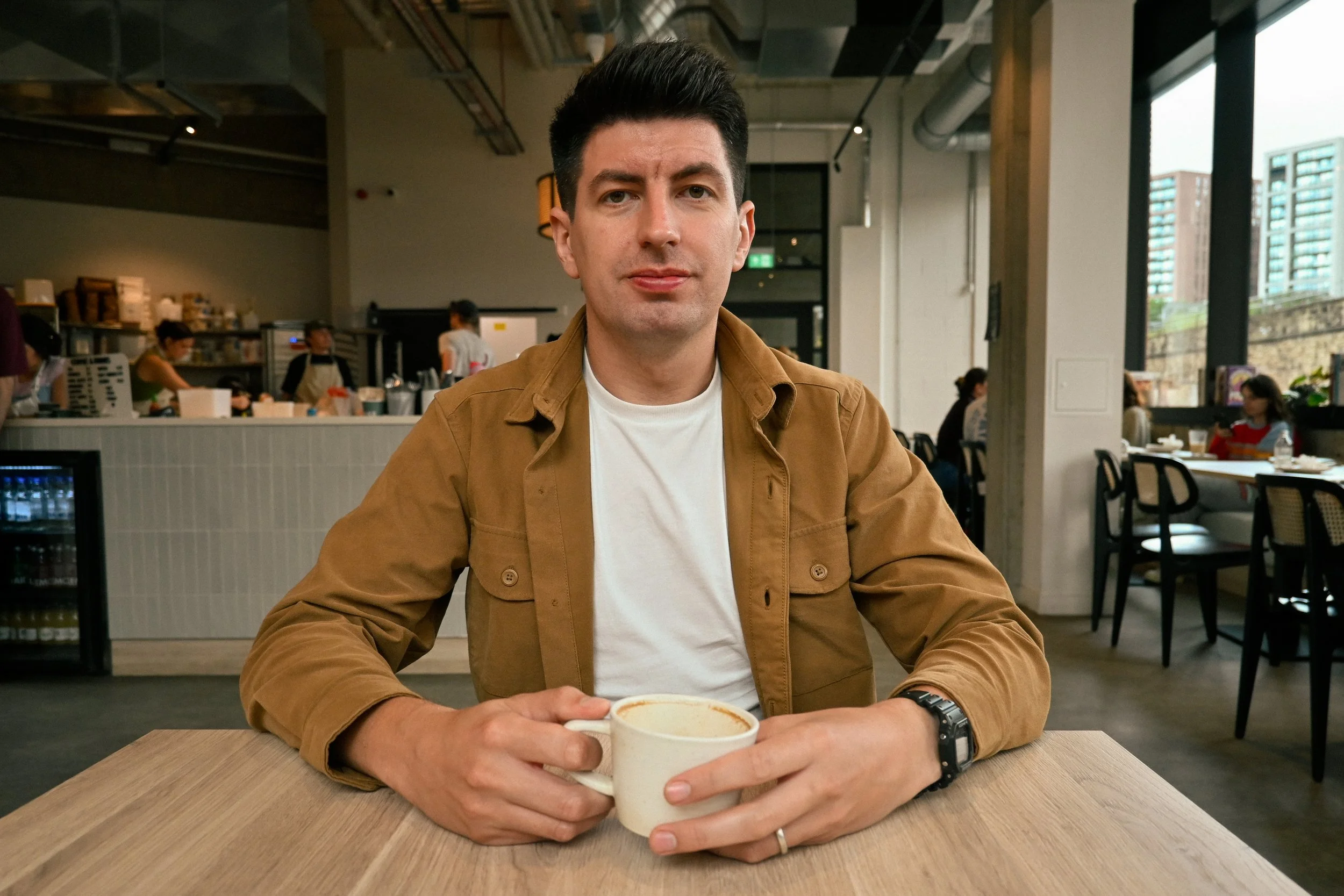 A man with dark hair wearing a brown jacket and white shirt sitting in a cafe, holding a cup of coffee, with other people and a counter in the background.