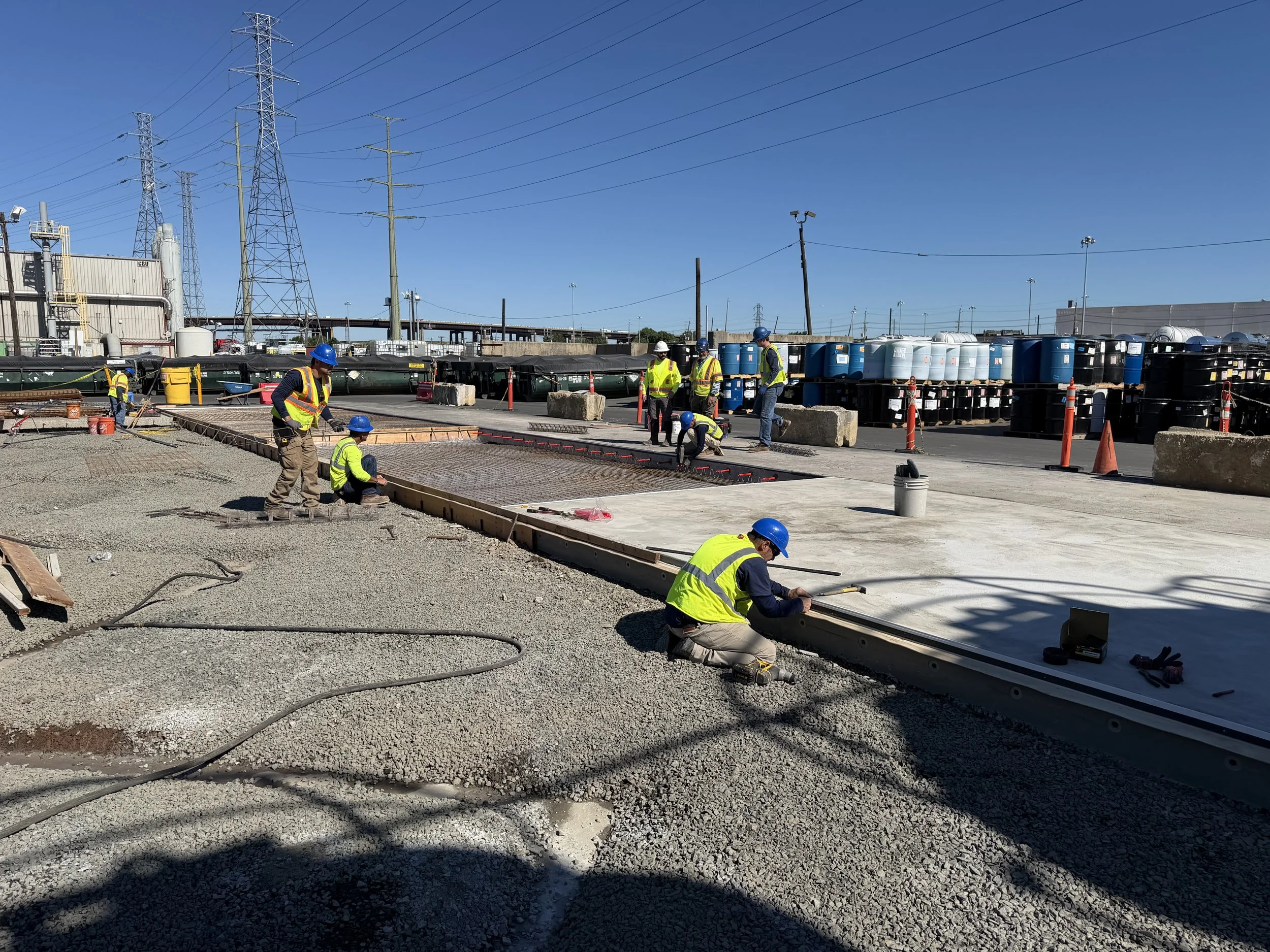 Workers in safety vests and helmets are laying concrete on a construction site with pipes, barrels, and power lines in the background.