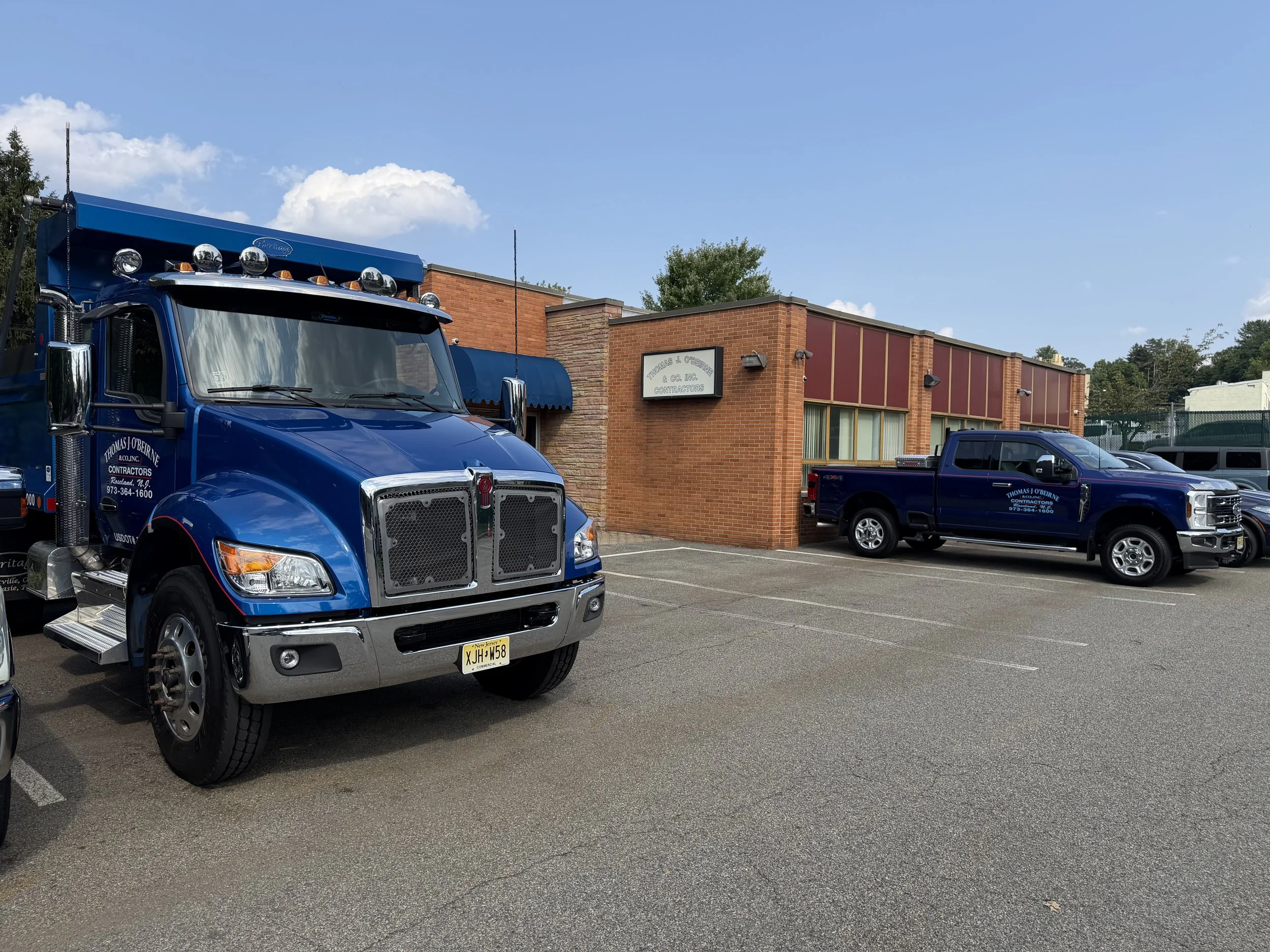 Parking lot scene with a large blue tow truck in the foreground. In the background, there is a brick building with business signage for Thomas J. O'Brien & Co. Inc., contractors. Several other vehicles are parked, and the sky is partly cloudy.