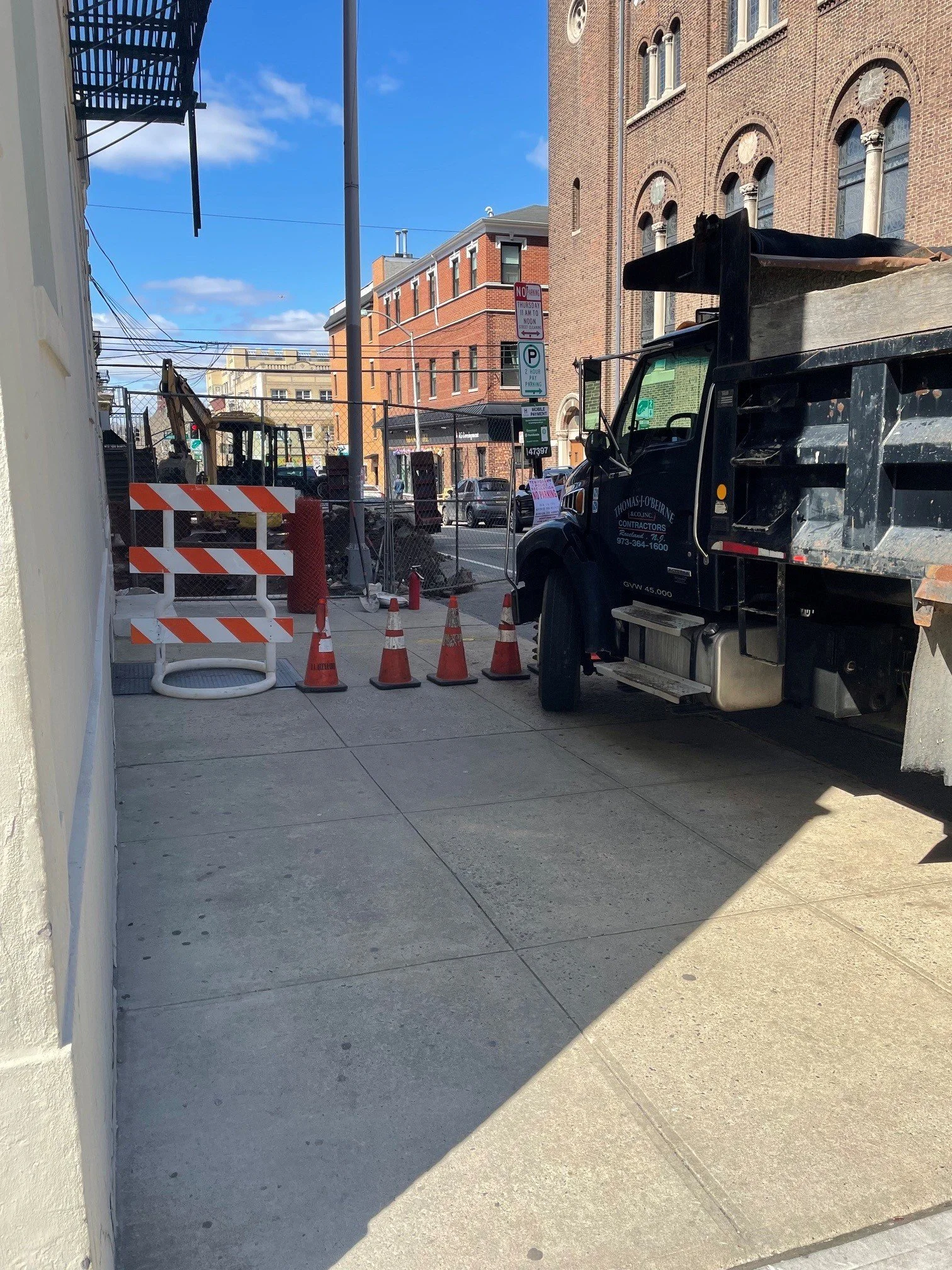 City street with construction zone, orange and white barricades, traffic cones, parked cars, brick buildings, and a blue sky with a few clouds.
