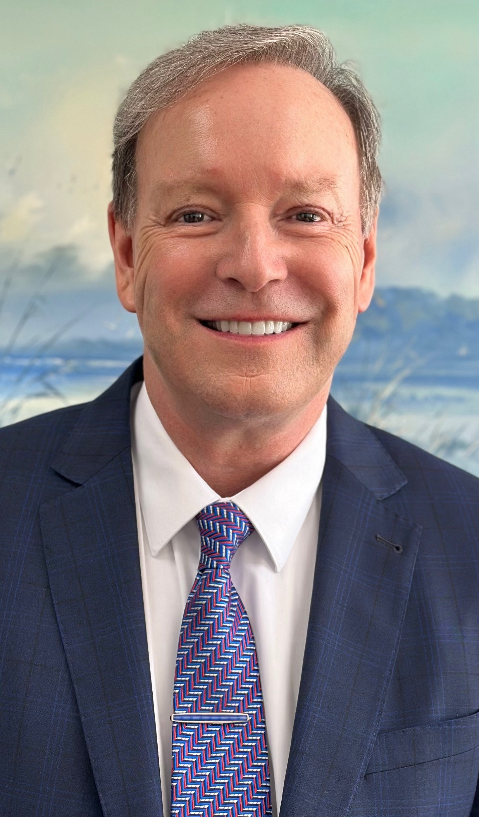Headshot of a smiling man in a business suit, blue checkered blazer, white shirt, and a colorful patterned tie, against a blurred scenic background.