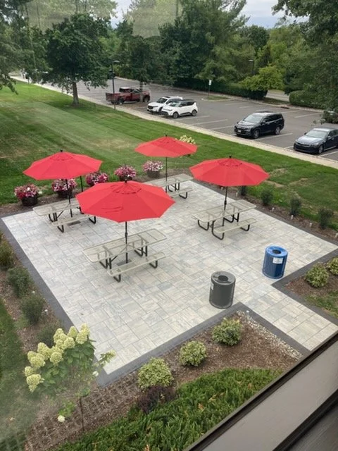 Empty outdoor patio with four red umbrellas over picnic tables, pink flowers in pots, two trash bins, surrounded by a lawn and parking lot.
