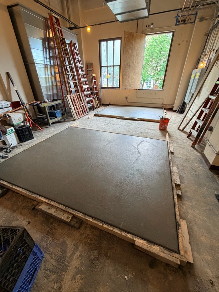 Indoor construction site with freshly poured concrete on a wooden frame, surrounded by ladders, tools, and large windows letting in natural light.
