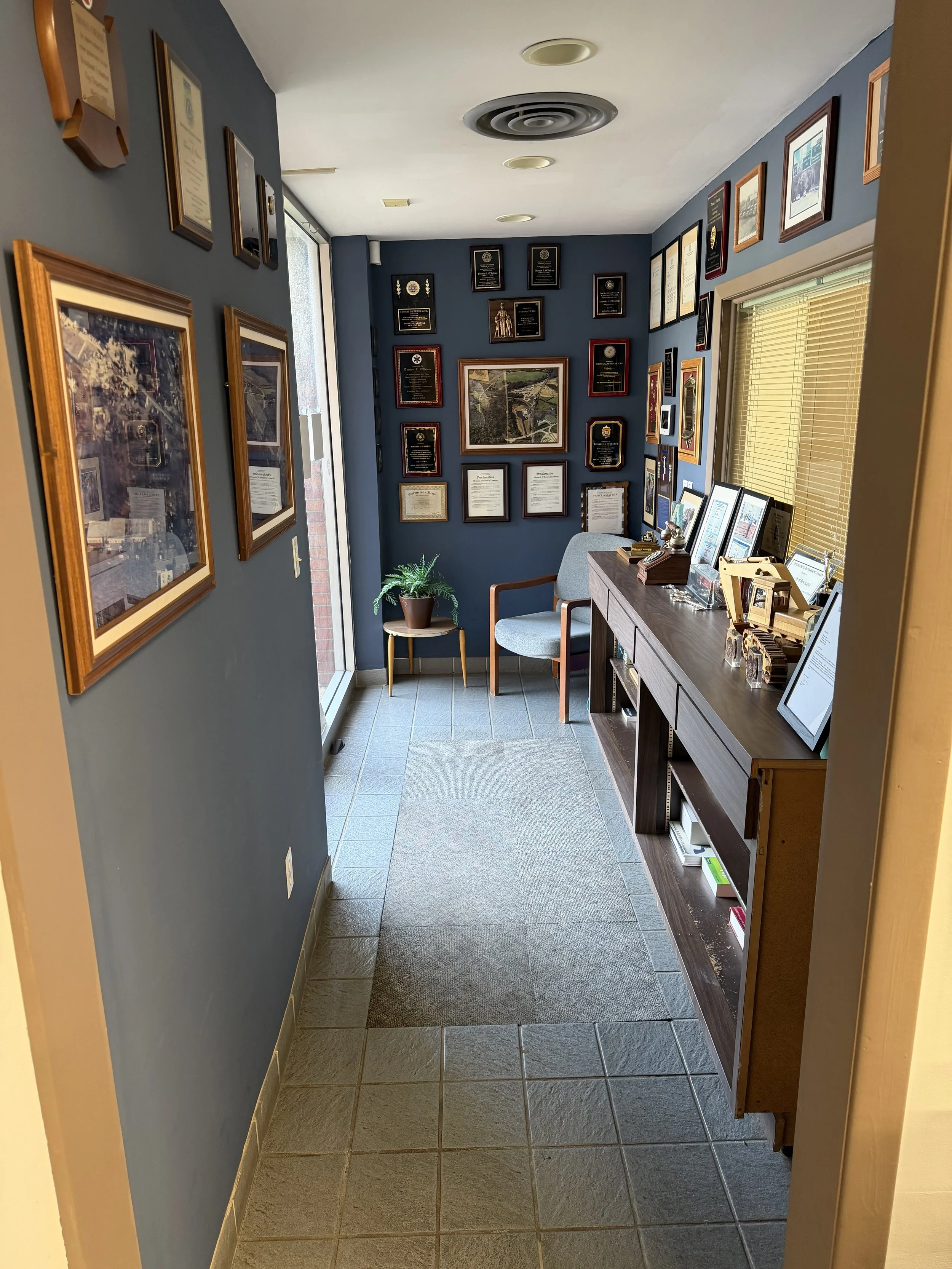 A small office or reception area with a blue wall decorated with framed awards and certificates. A gray upholstered chair and a small table with a potted plant are near a window with closed blinds. A wooden sideboard on the right holds framed documents and decorative items. The floor is tiled, and ceiling lights are visible.