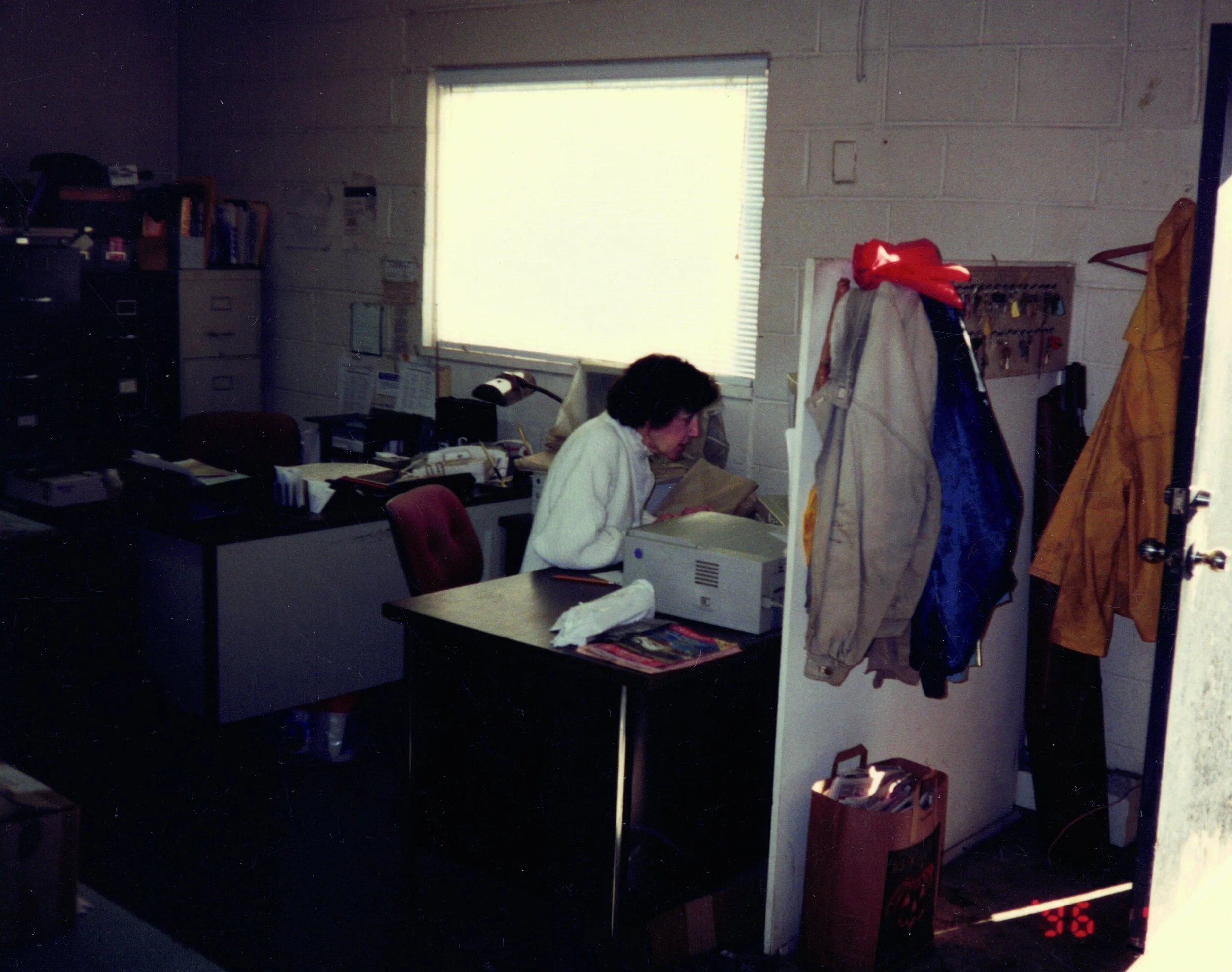 Office space with a person working at a desk, surrounded by filing cabinets, office supplies, and clothes hanging on a rack, near a window with blinds.