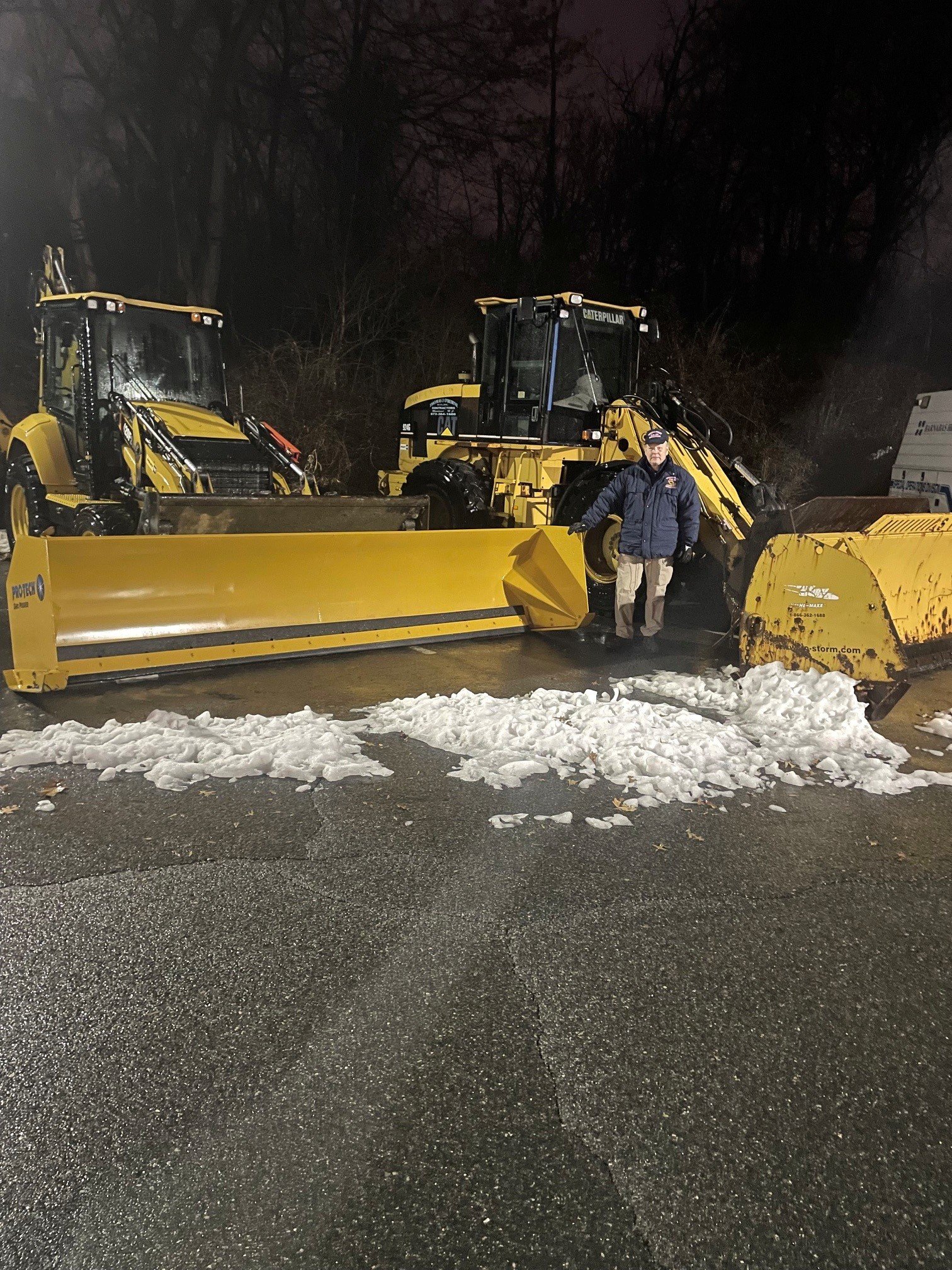 A man standing in front of large yellow snow plows on a wet road at night, with snow on the ground and leafless trees in the background.