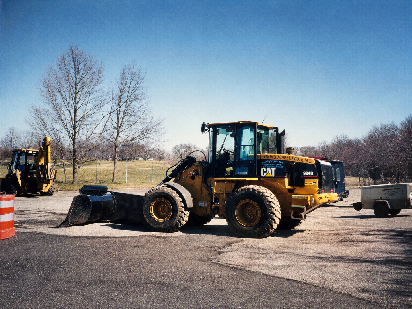 Construction site with a large yellow CAT 924G wheel loader, a smaller backhoe loader, an orange traffic barrel, and a trailer. Bare trees and a blue sky in the background.