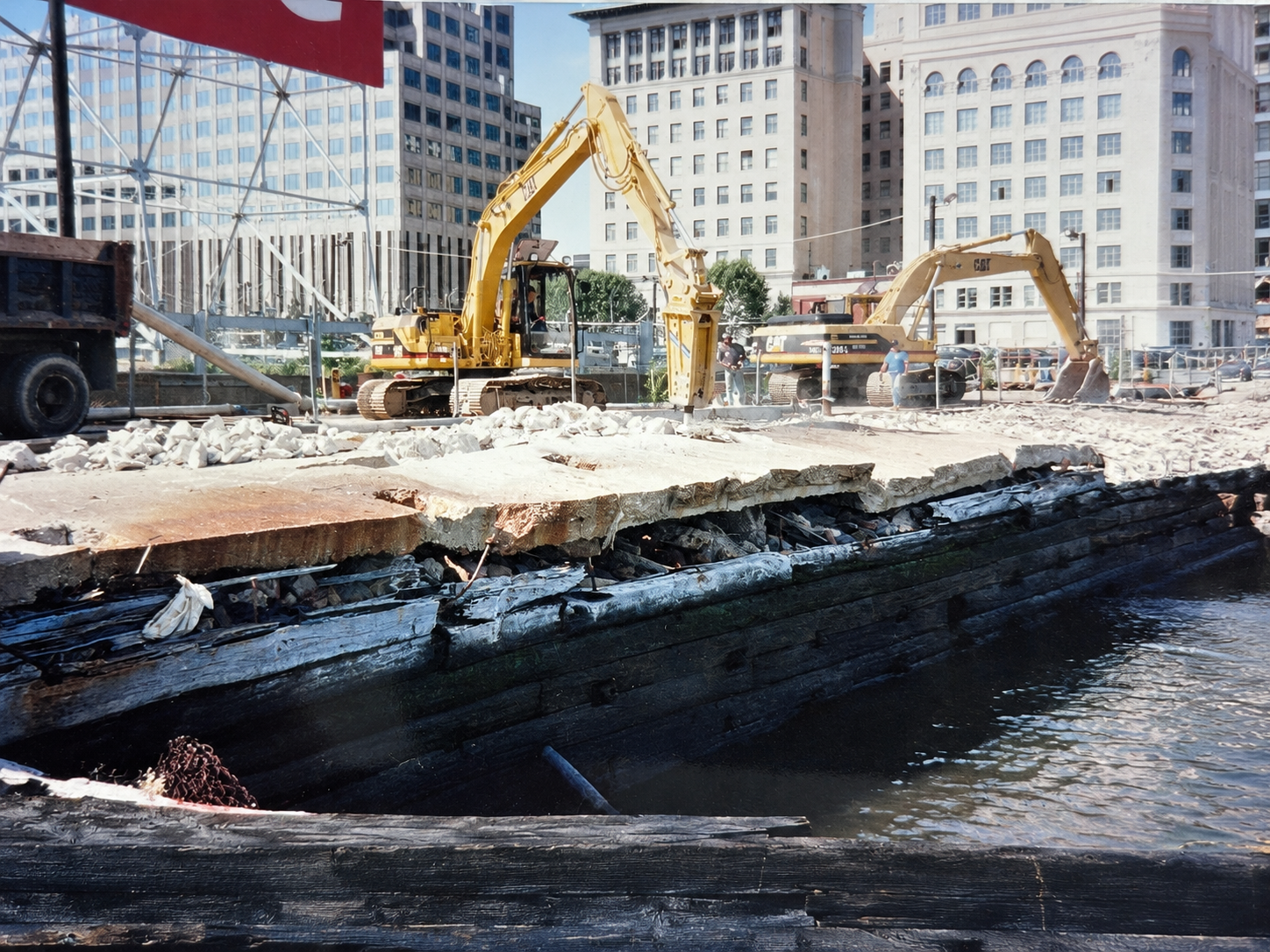 Construction site near water showing two large yellow excavators, construction workers, concrete debris, and tall buildings in the background.