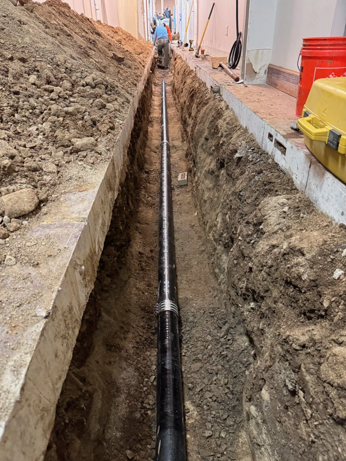 Construction workers installing a pipe in a long, narrow trench inside a building.