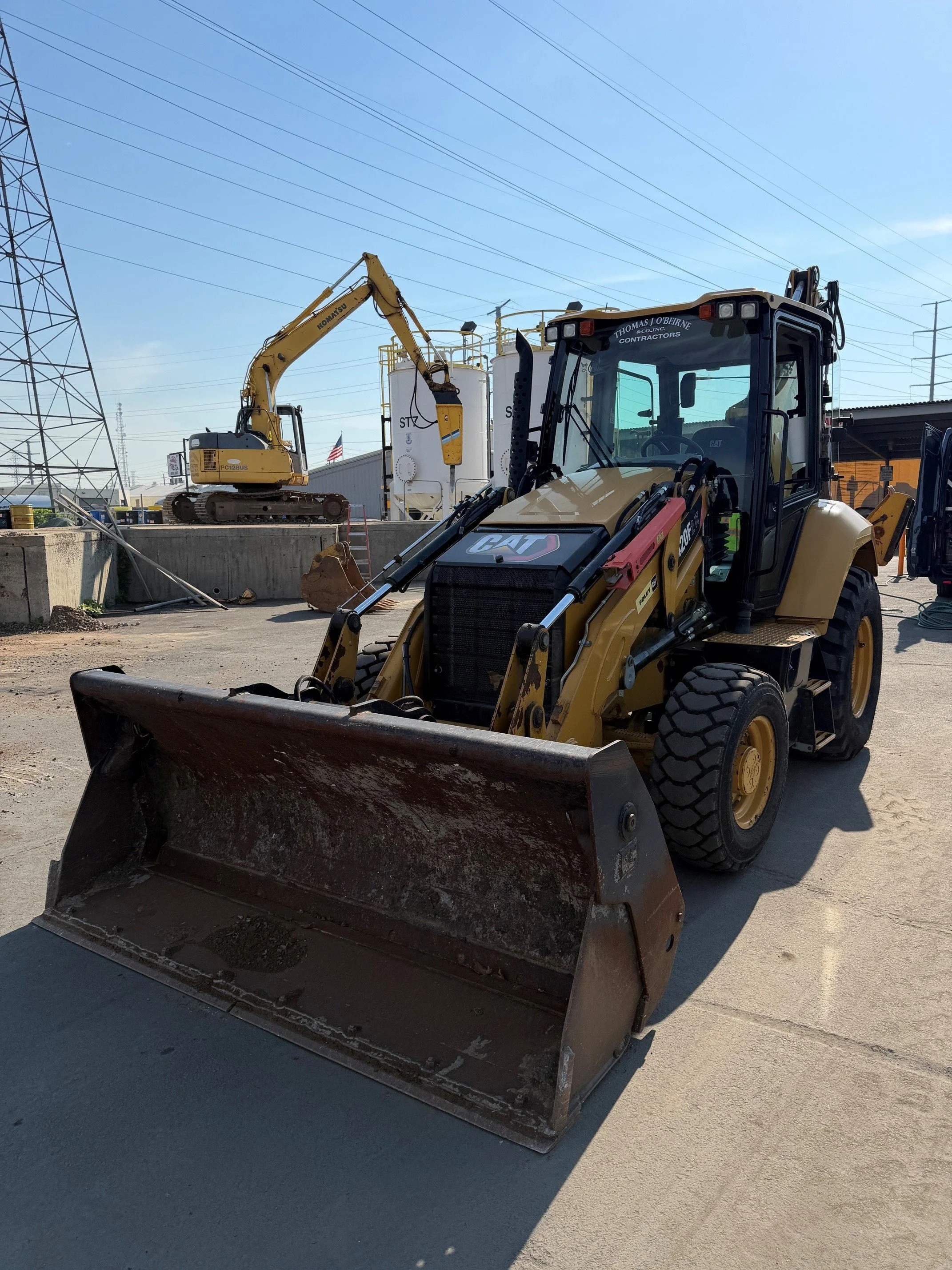 Construction site with a front loader and an excavator, power lines overhead, and a large storage tank in the background.