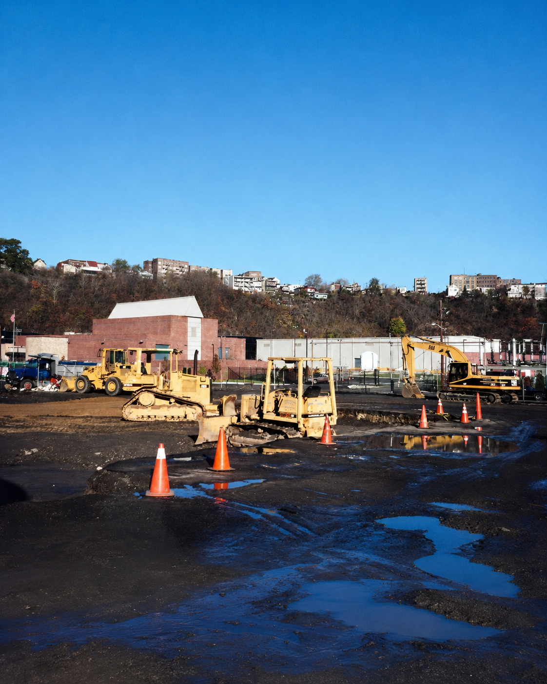 Construction site with bulldozers and orange safety cones on muddy ground, with a hill and buildings in the background under a clear blue sky.