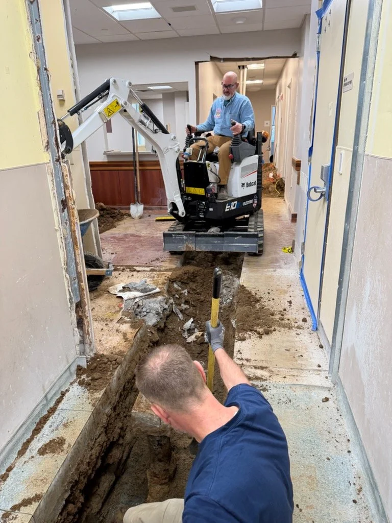 Construction workers lift flooring, with one using a small excavator to remove the floor, and another worker digging a trench with a shovel in an indoor building under renovation.