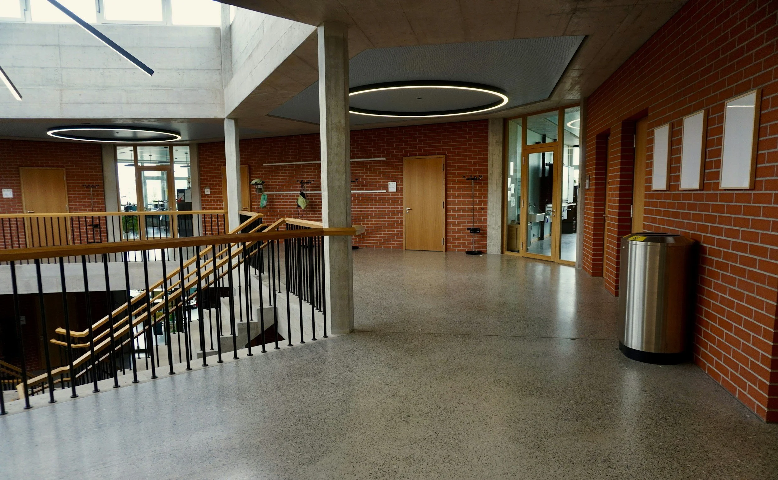 Interior view of a modern building with brick walls, a staircase with wooden railings, circular ceiling lights, and glass doors.