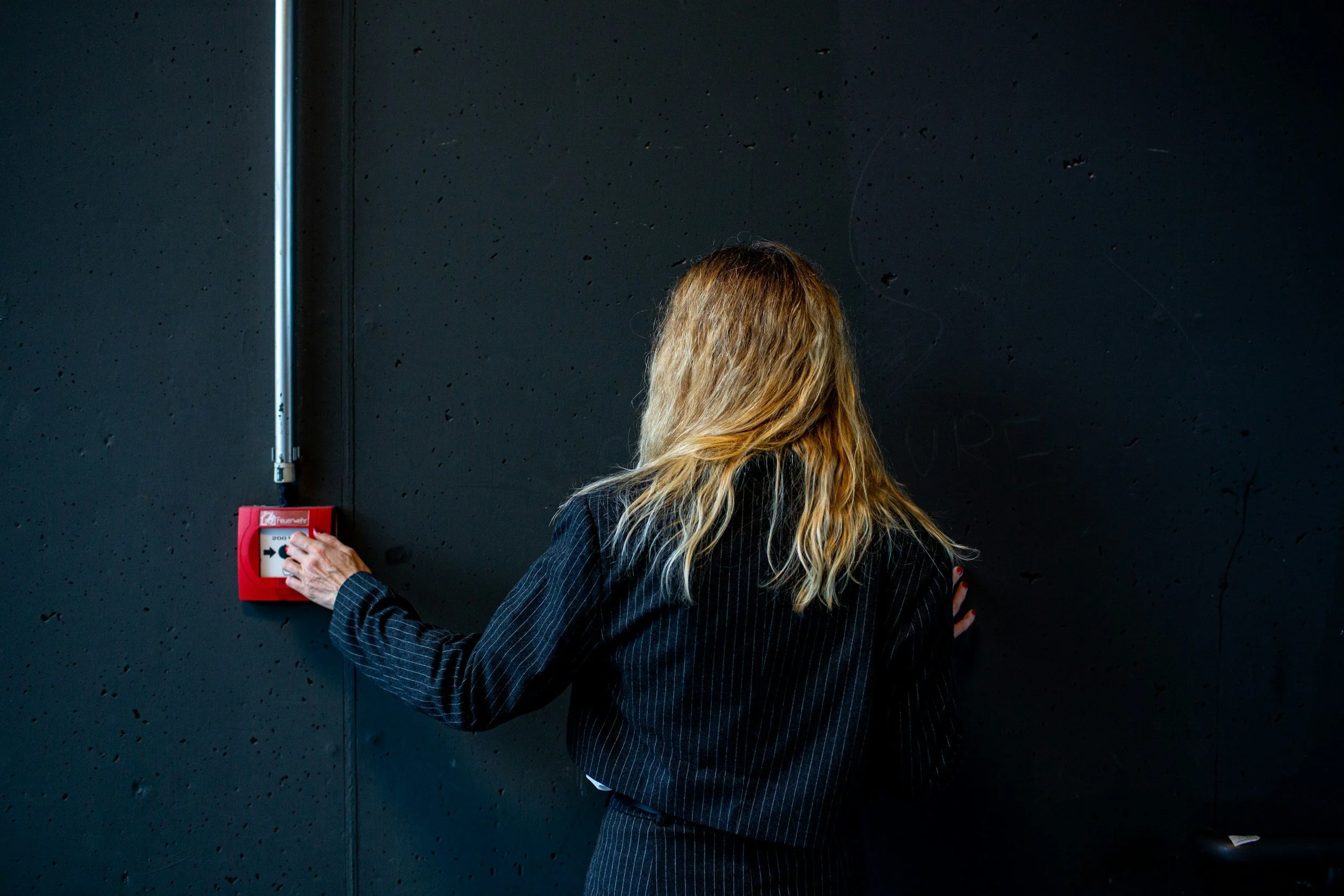 A woman with long blonde hair, dressed in a dark pinstripe suit, is pressing a button on a red fire alarm pull station mounted on a black wall.