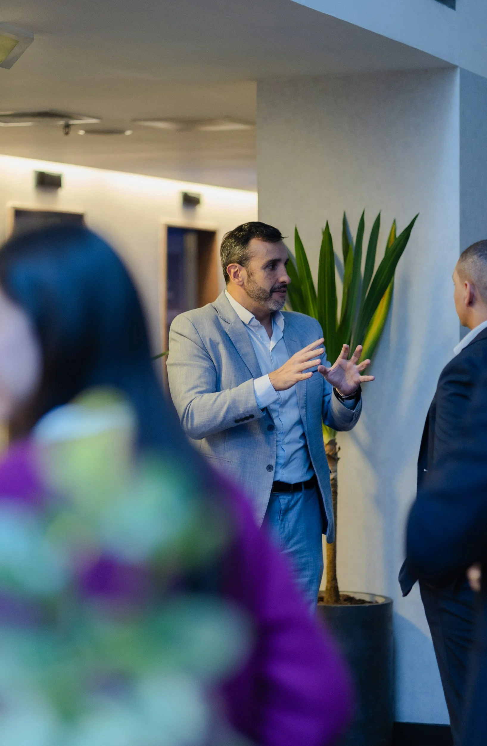 Strategic Estate Management Training for Schools. Man in a light gray suit and white shirt is speaking and gesturing with his hands to a person in a dark suit in an indoor setting. There is a large green plant and a blurred person in the foreground.