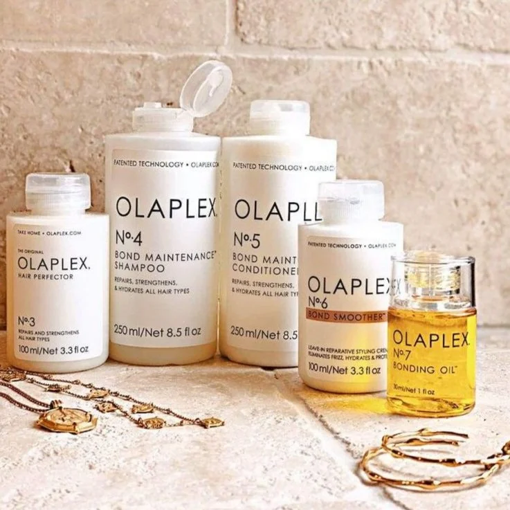 A collection of Olaplex hair care products, including shampoo, conditioner, styling cream, and bond oil, arranged on a light-colored surface with jewelry, in front of a beige brick wall.