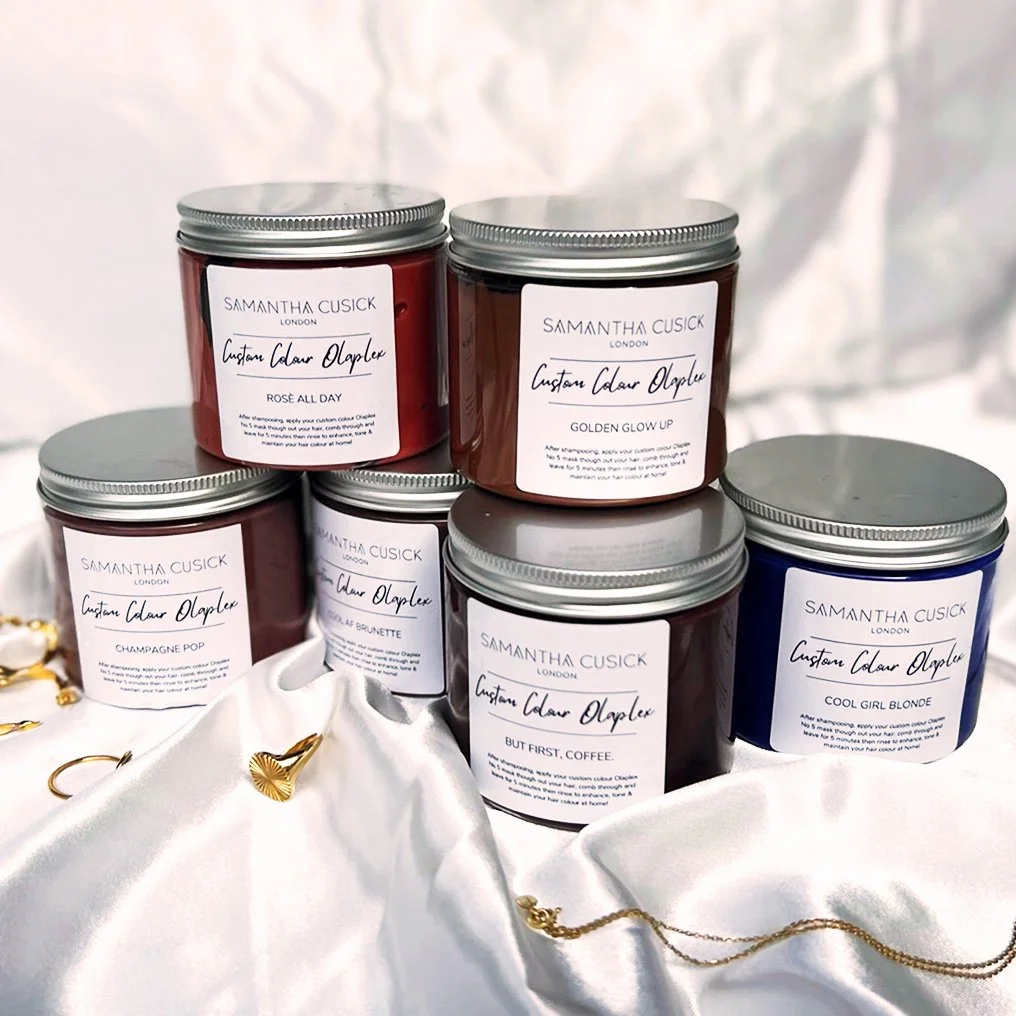 Seven scented candles in glass jars with metal lids, labeled with various names and scents, placed on white fabric with small gold jewelry pieces.