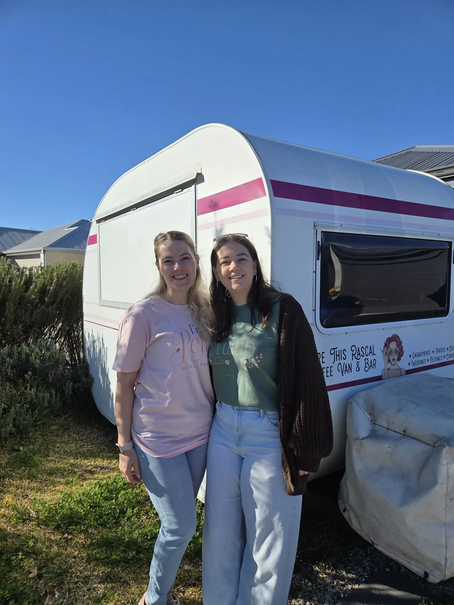 Two women standing outdoors in front of a white trailer with pink stripes, smiling at the camera on a sunny day with a blue sky overhead.