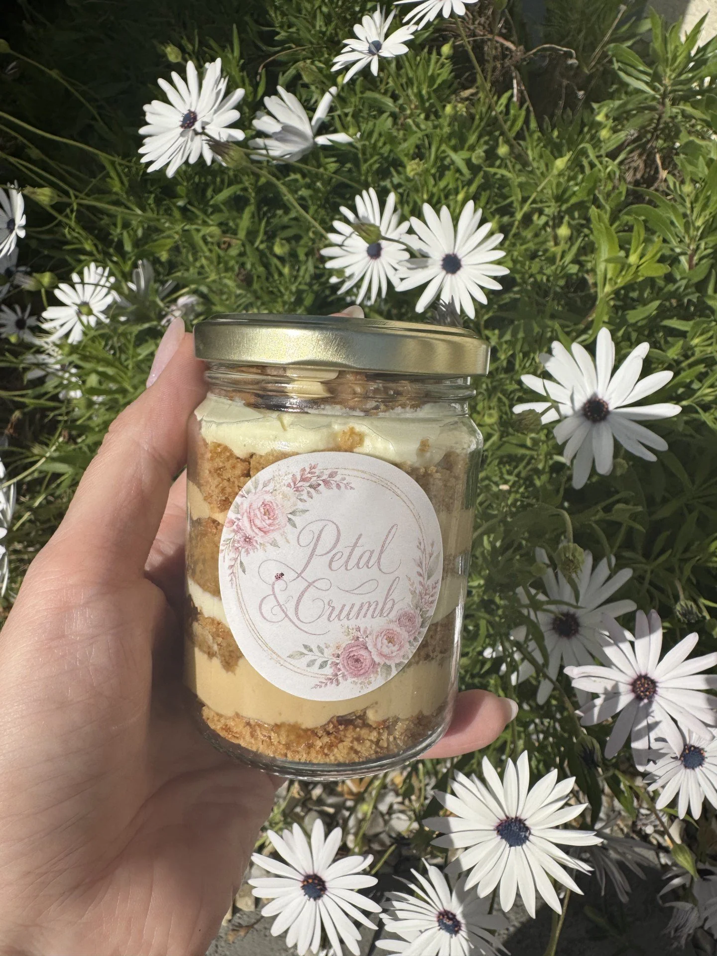 A jar of Petal & Crumb cookie crumbs held in front of white daisies with dark centers and green foliage.