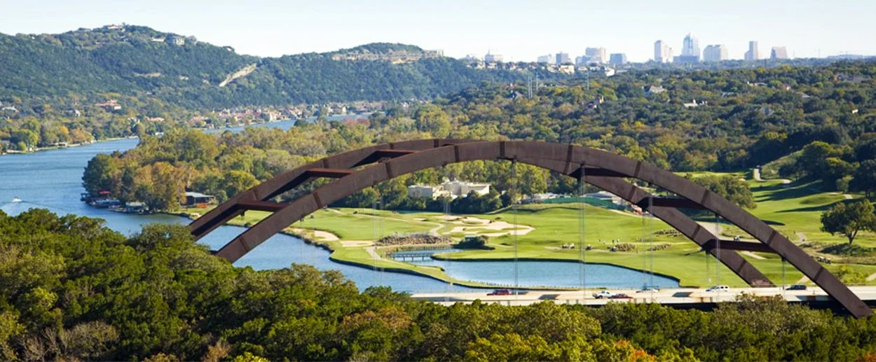 Large bridge with a curved arch over a river, surrounded by green trees and a golf course, with a city skyline in the distance.