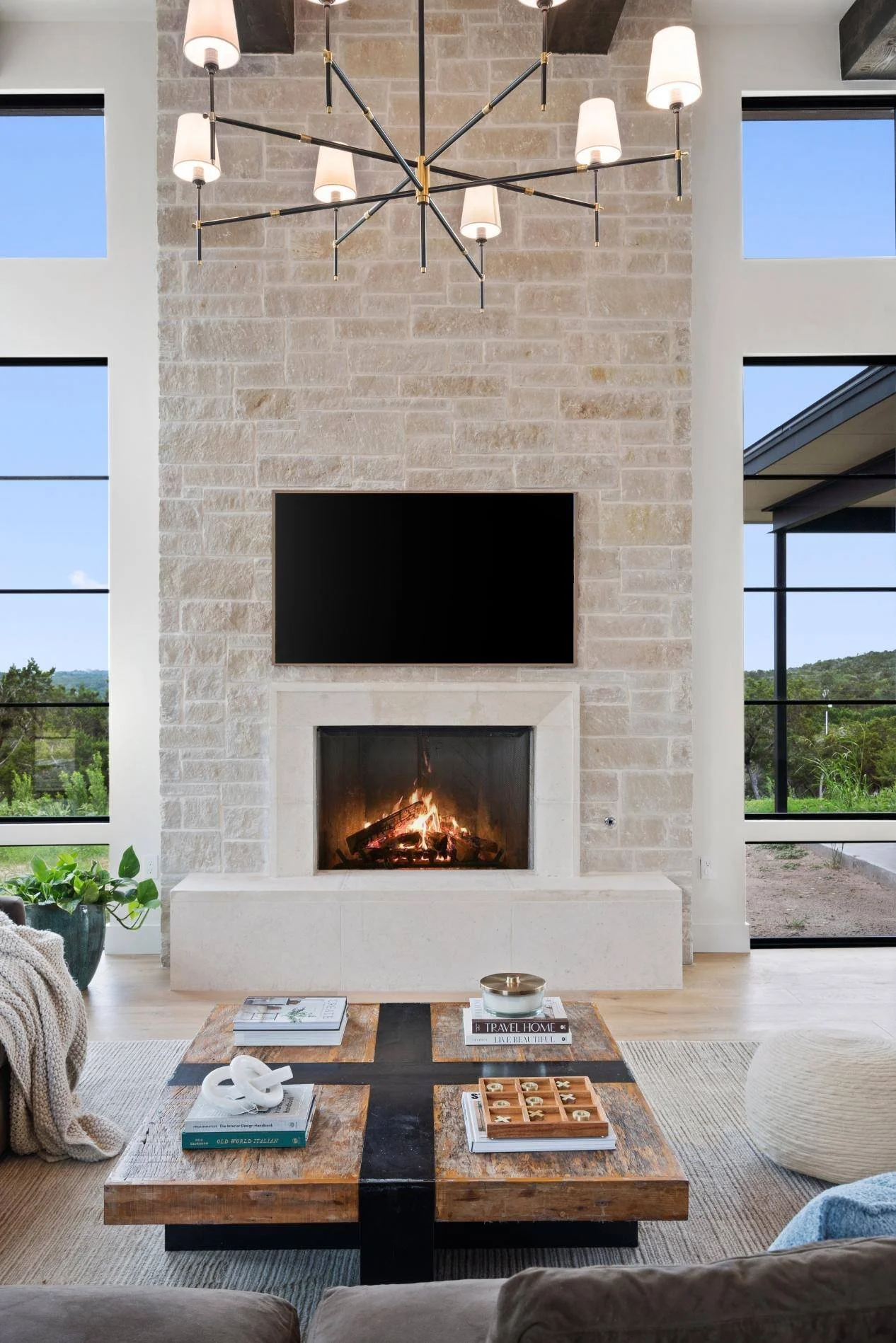 Living room with a modern fireplace, a mounted flat-screen TV above it, a large wooden coffee table with books, a coffee cup, and decorative items, beige sofa, and large windows showing a green outdoor landscape.