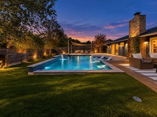 Backyard with illuminated swimming pool, outdoor seating, trees, and a house with a chimney at dusk.