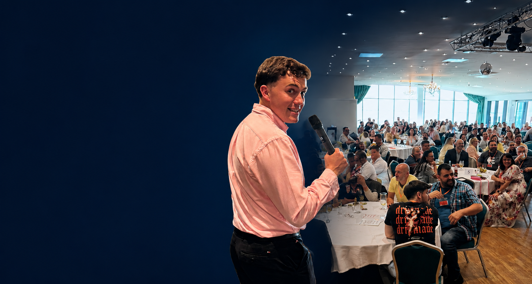 Man holding a microphone standing in front of a large seated audience in a banquet hall with windows and chandeliers.