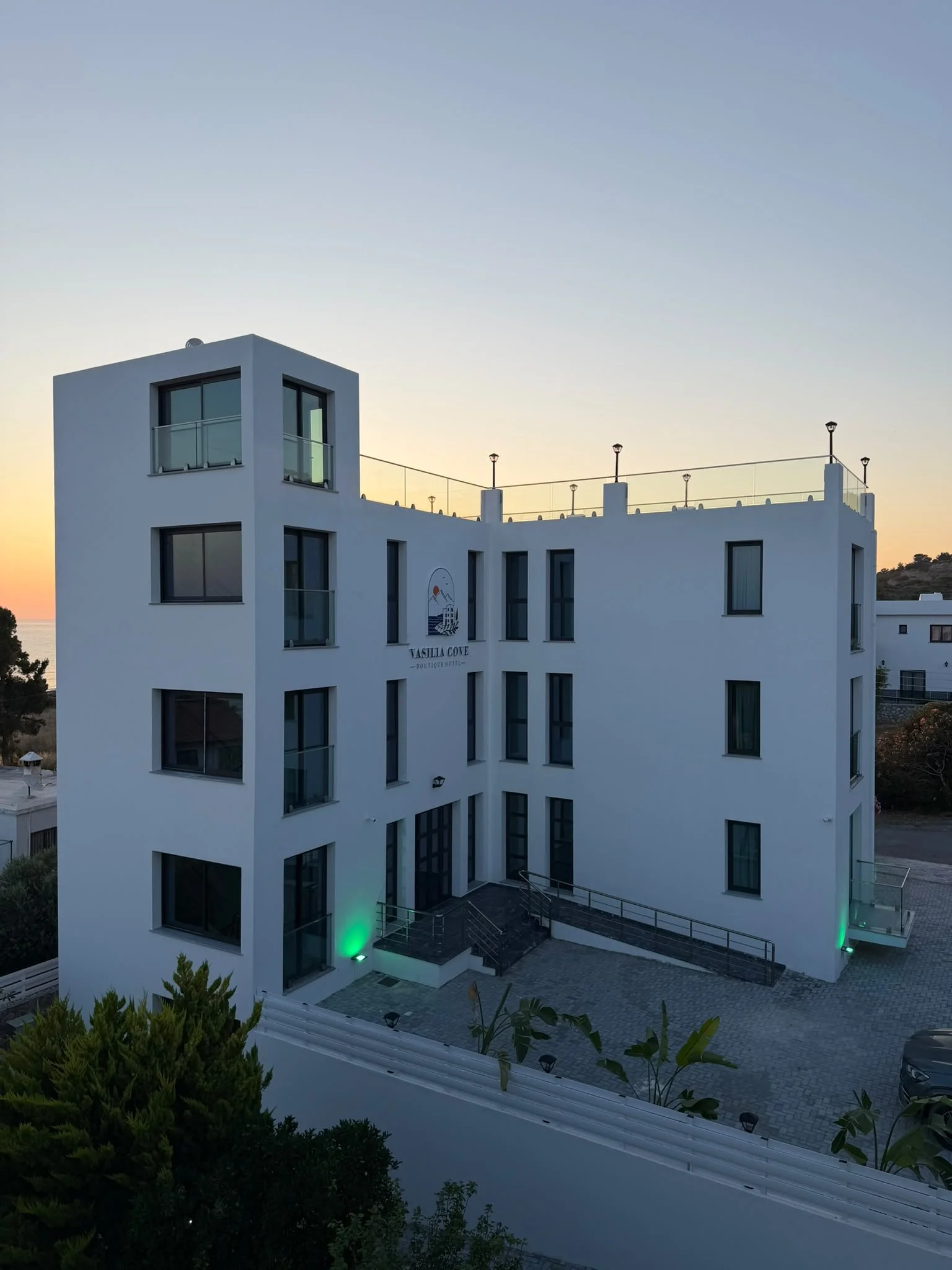 Modern white multi-story building with large glass windows at sunset, green lighting at the entrance, and a balcony on the top floor with railings and several outdoor lights.