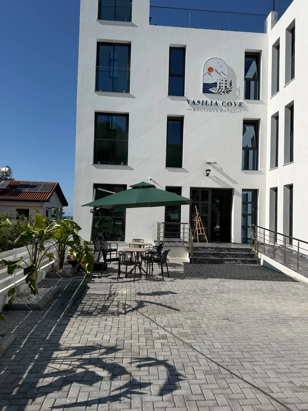 Exterior of the Vasila Cove Boutique Hotel with outdoor seating area, a green umbrella, black chairs, and potted plants, under a clear blue sky.