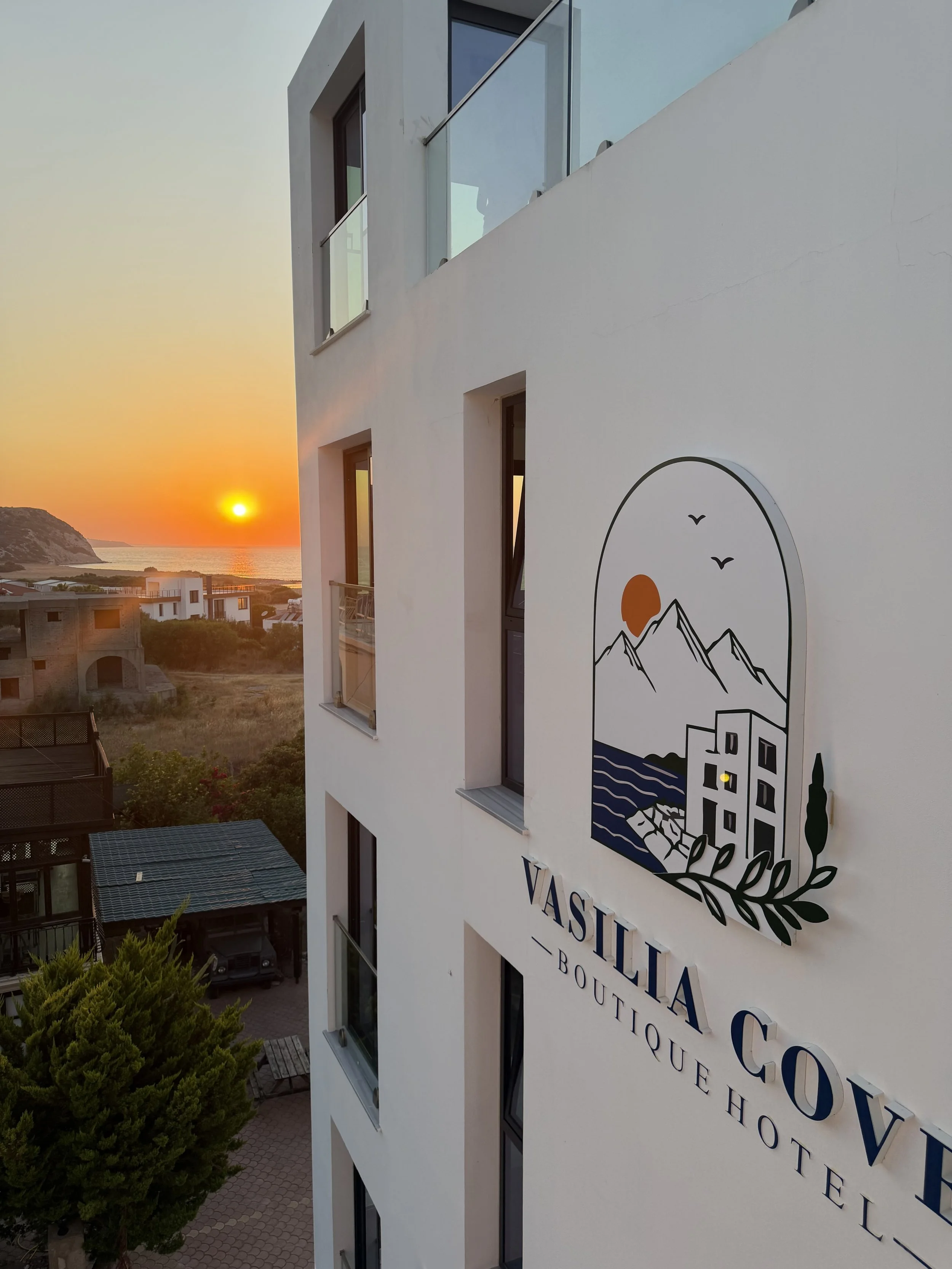 View of a white modern building with a logo and partly visible sign, overlooking the sunset over a coastal area with hills and water.