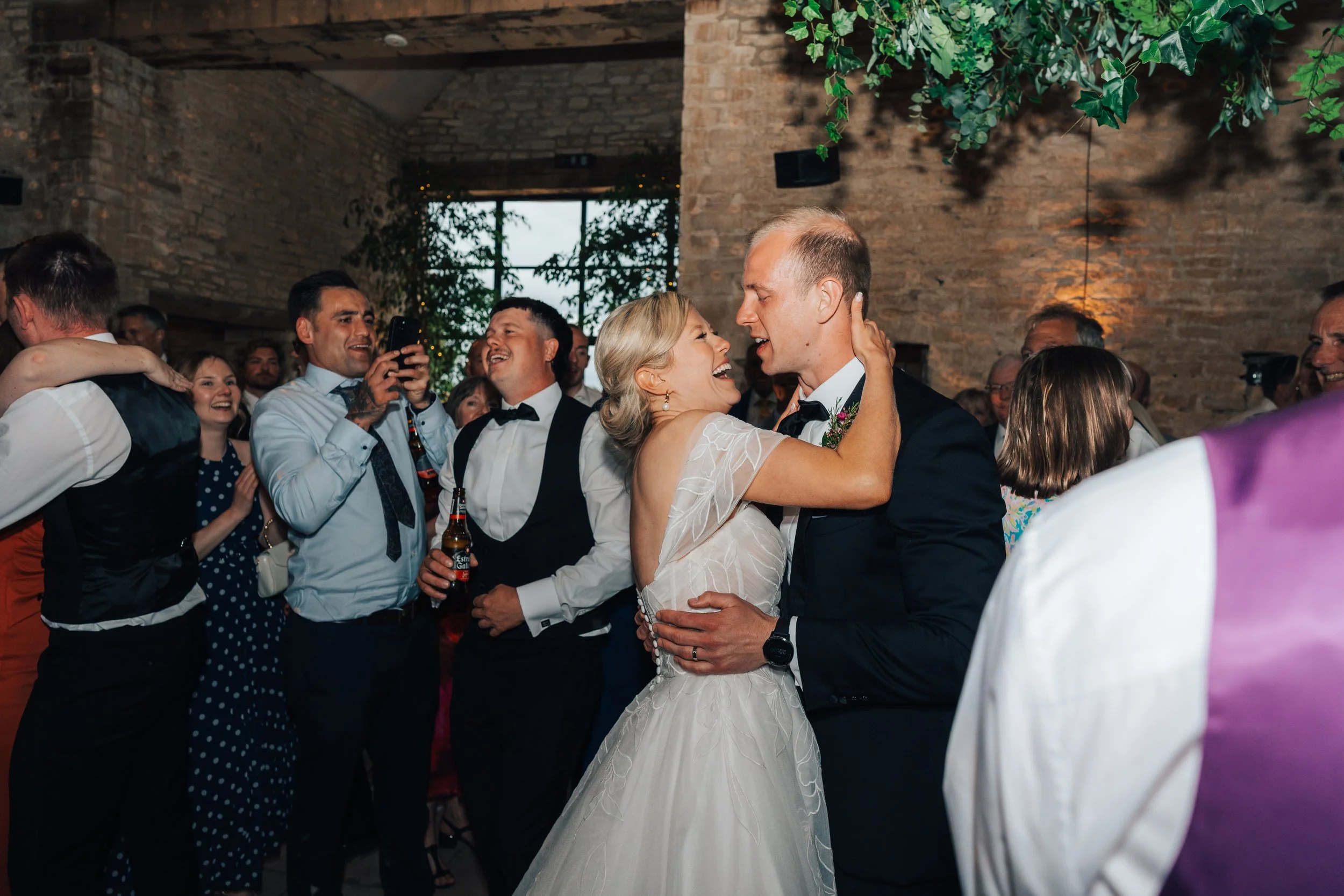 A bride and groom dance closely during their wedding reception, surrounded by happy guests in an indoor venue with brick walls and greenery.