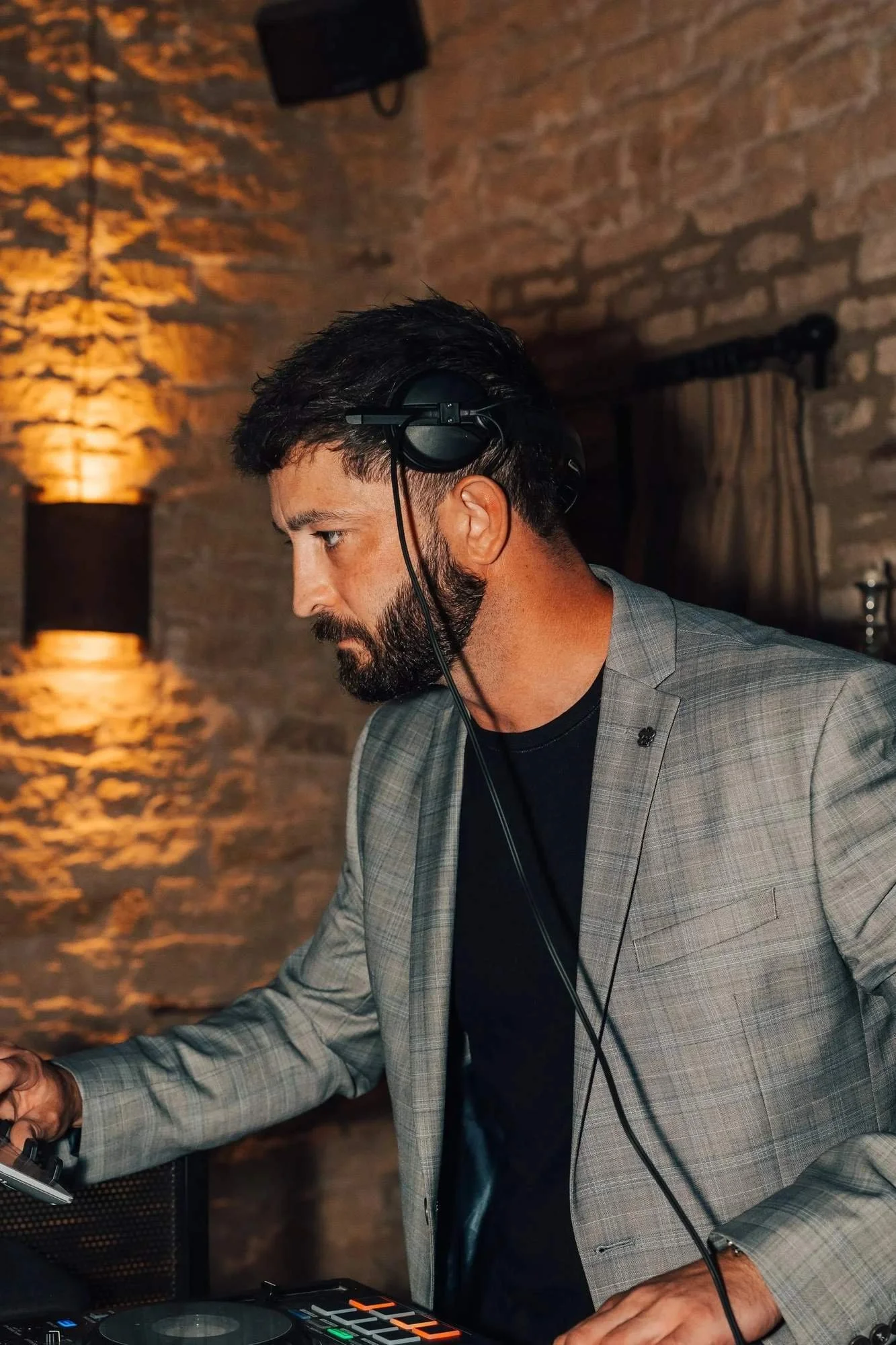A man with dark hair, a beard, and light skin wearing a gray checkered blazer and black shirt, DJing with headphones on in a dimly lit rustic venue with brick walls and warm lighting.