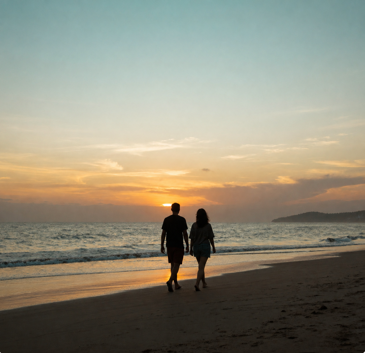 A couple walking hand in hand along the beach at sunset, with the ocean and a distant landform in the background.