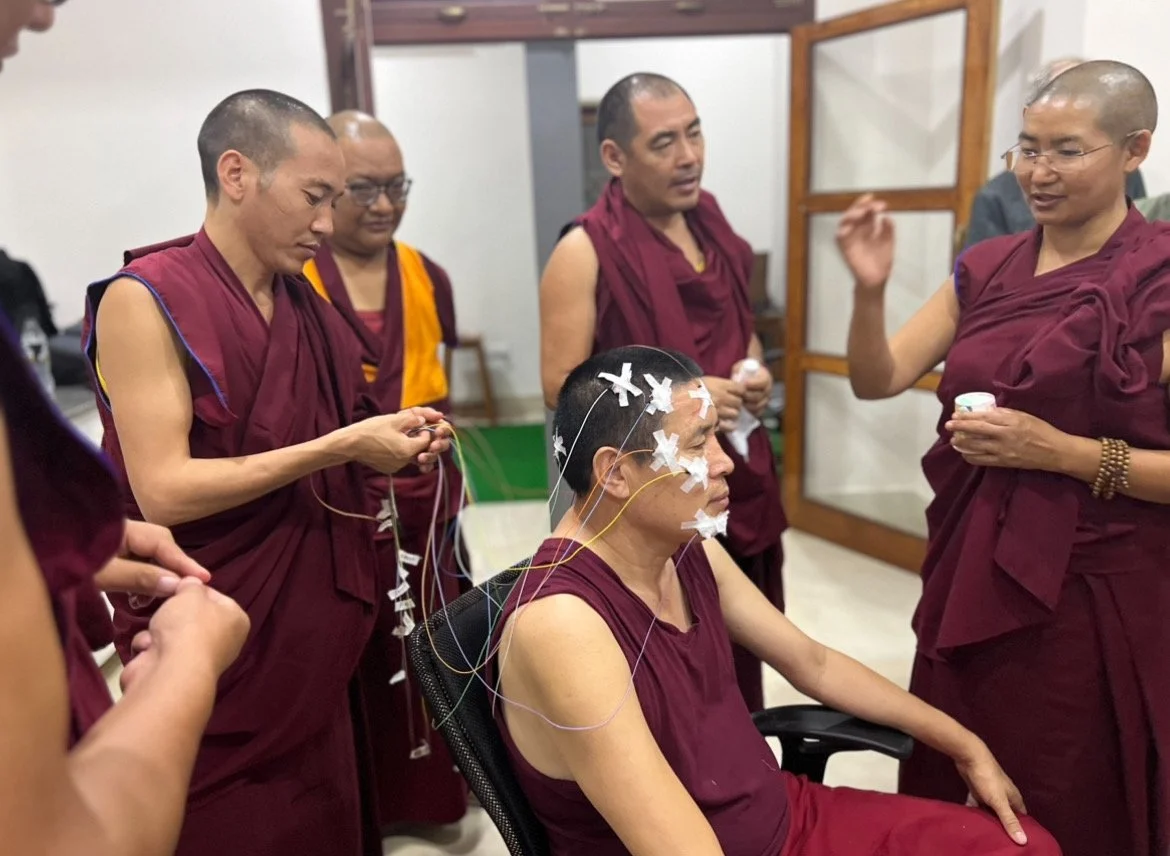 Buddhist monks observe a seated man undergoing an EEG test with electrodes attached to his face and scalp.