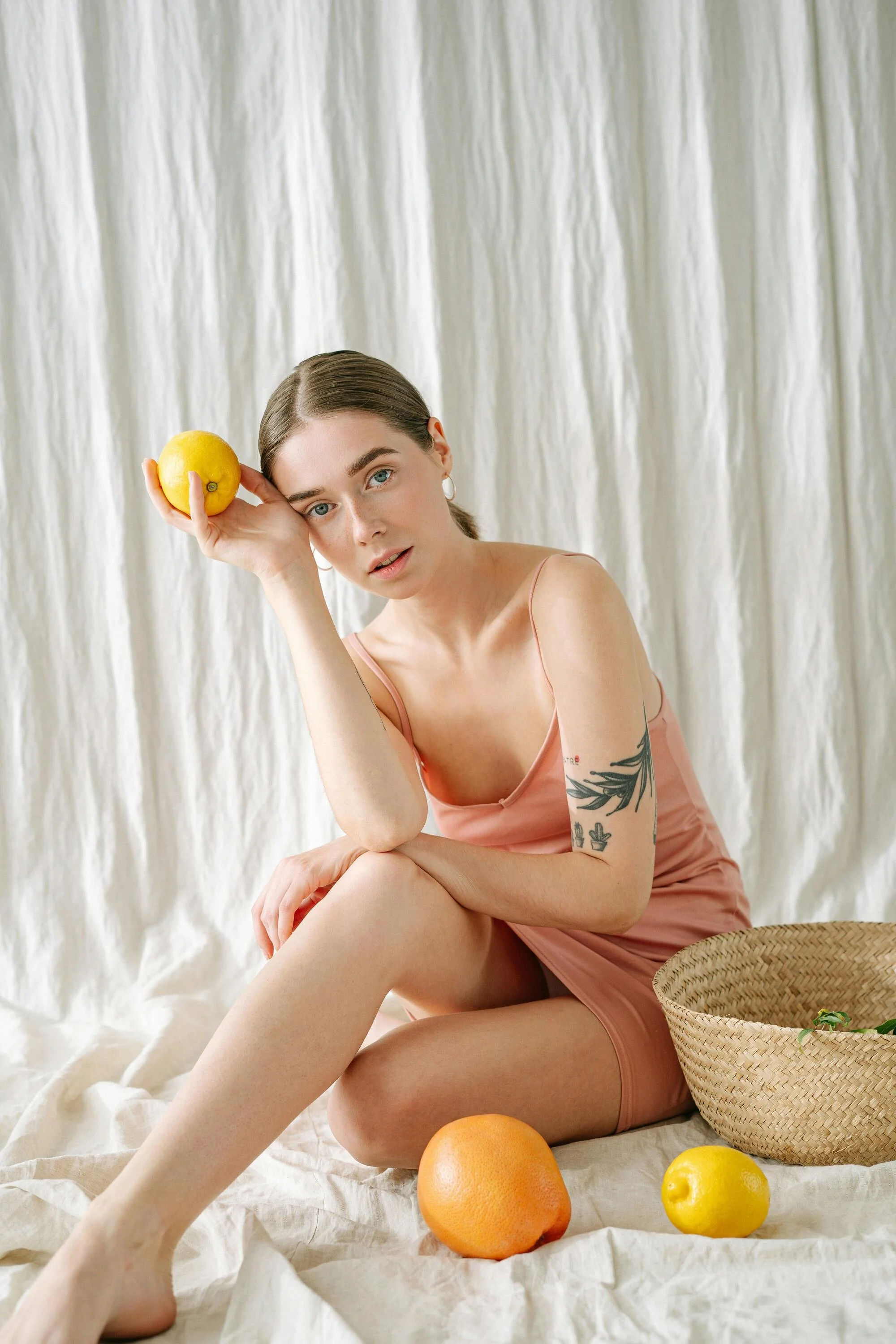A young woman with blue eyes and tattoos on her arm, wearing a peach-colored spaghetti strap dress, sitting on a white fabric backdrop with a basket and assorted citrus fruits including oranges and lemons around her, holding a lemon near her temple with a neutral expression.