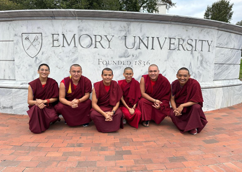 Six people dressed in maroon Buddhist robes sitting on brick pavement in front of a white marble sign reading 'Emory University Founded 1836' with a shield emblem.