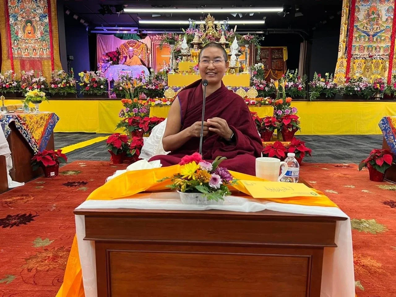 A smiling monk in maroon robes sitting behind a table decorated with flowers, with a microphone in front of her, in a hall decorated with colorful flowers and religious banners.