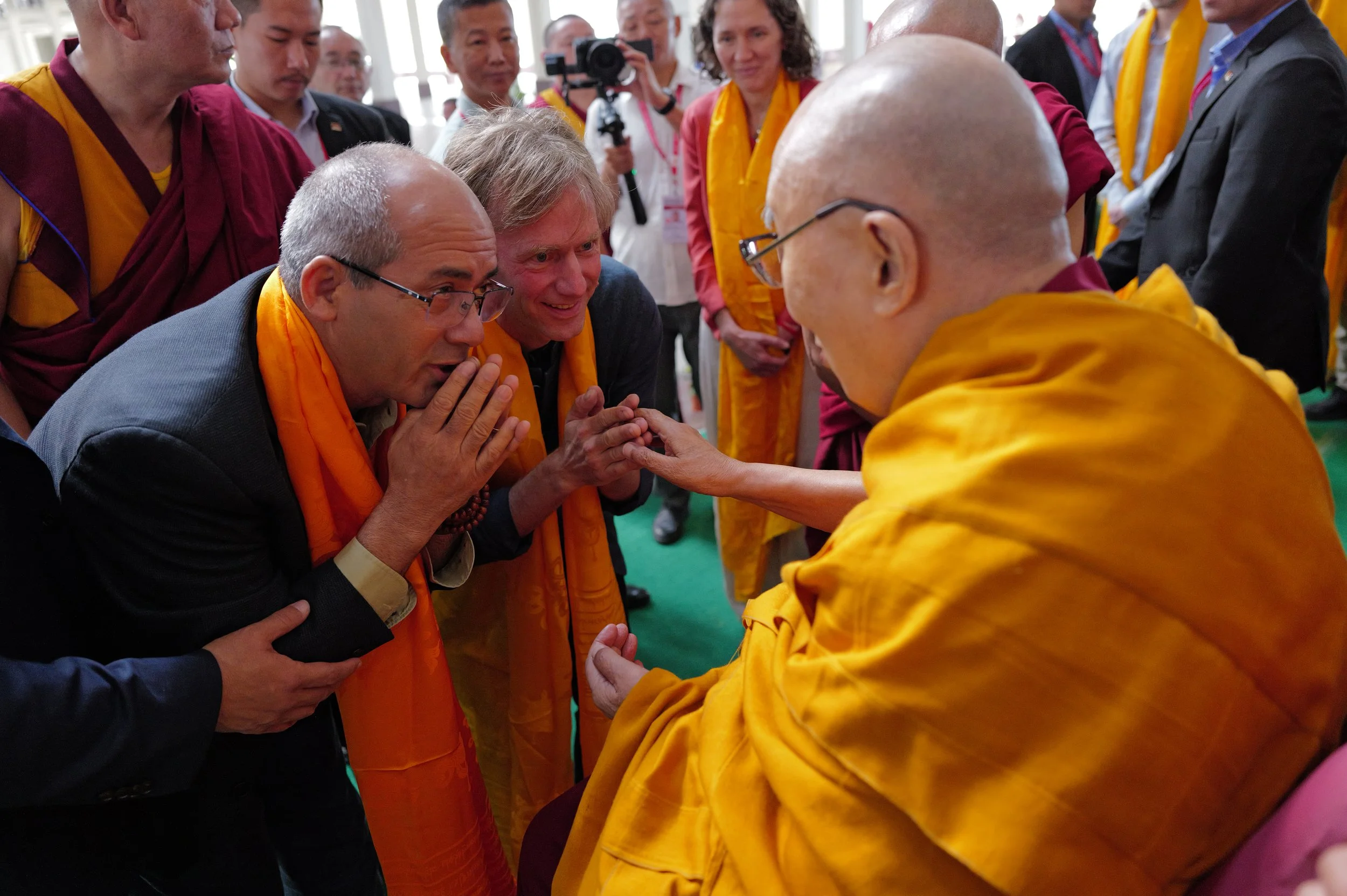 Buddhist monk in saffron robes greets two men kneeling respectfully at a crowded indoor event.