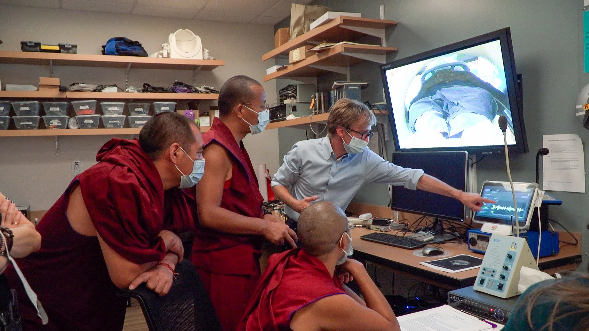 Group of medical professionals, including monks in traditional robes and a man in a shirt, gathered around medical equipment and a monitor in a clinical setting. One person is pointing at a screen showing an image from a camera, with other people obs