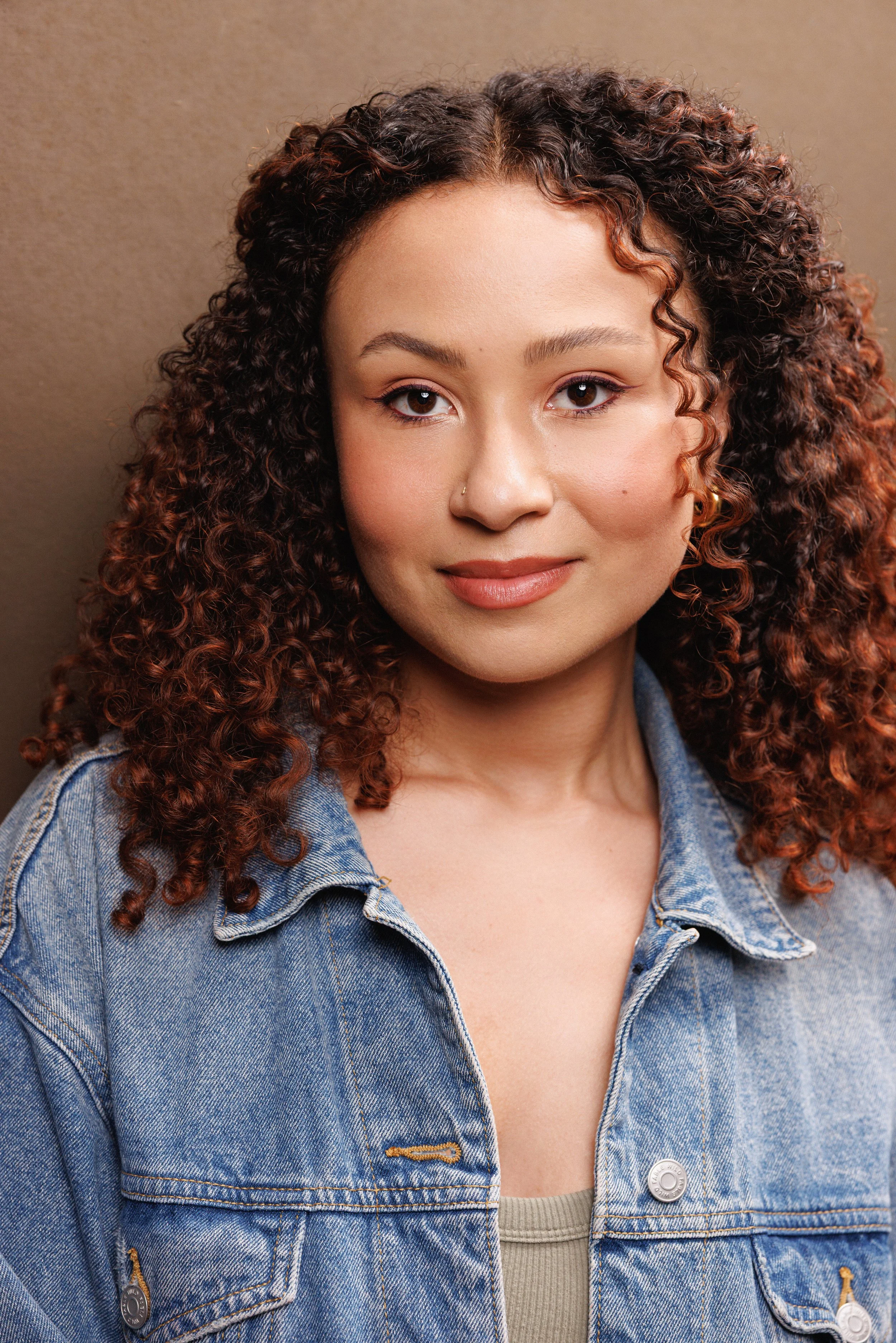 Close-up portrait of a smiling woman with curly dark brown hair, brown eyes, nose ring, and denim jacket