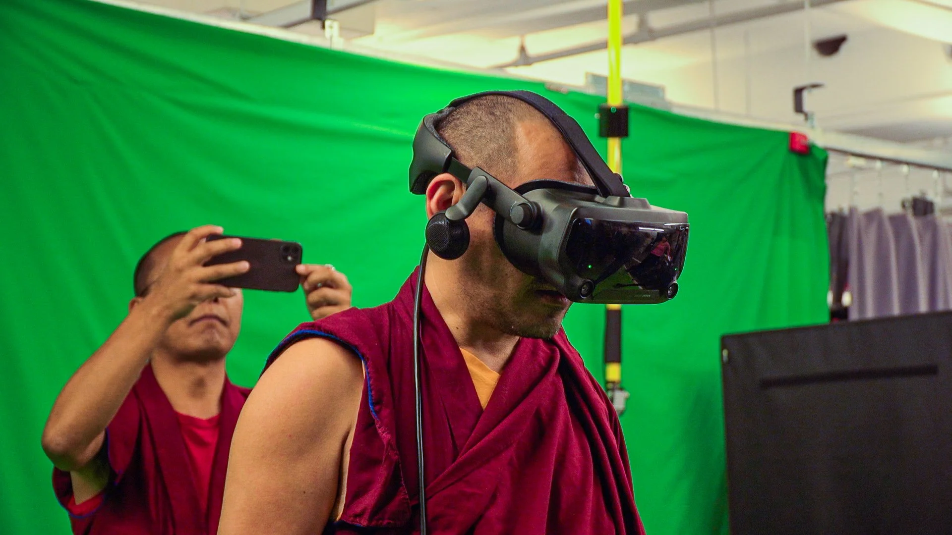 A person wearing virtual reality goggles, possibly a monk or athlete, in front of a green screen with another person taking a photo in the background.
