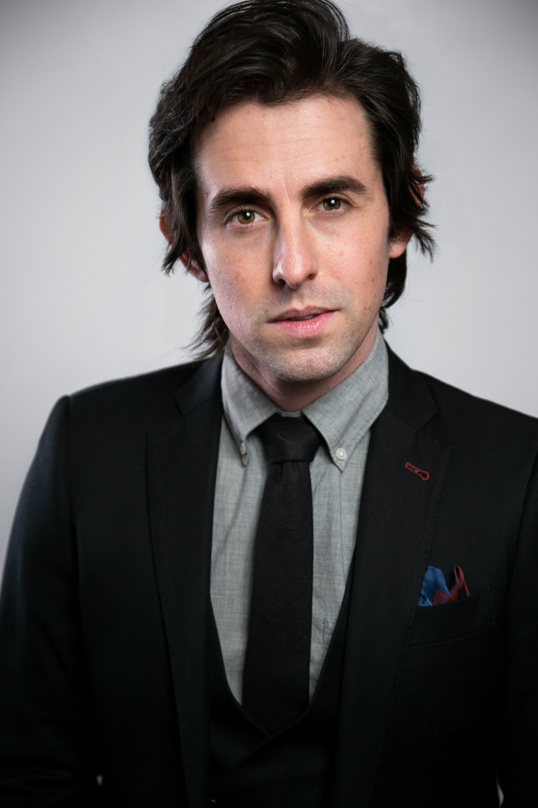 A portrait of a young man with dark brown, slightly wavy hair, wearing a dark suit jacket, gray dress shirt, and dark tie, with a pocket square, against a plain light background.